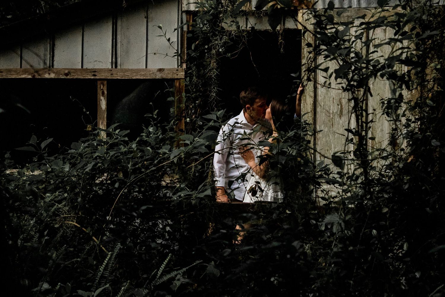 Couple sharing an intimate moment in a weathered doorway, framed by overgrown foliage. Moody, low-key lighting creates contrast, enhancing t