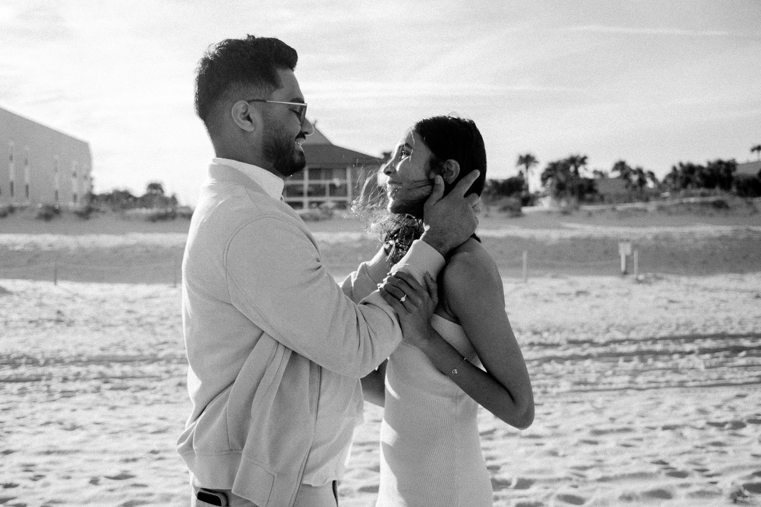 Engagement shoot on the beach, with the guy holding his fiancée’s head in his hands, looking at her in awe as she smiles and grabs his arms.