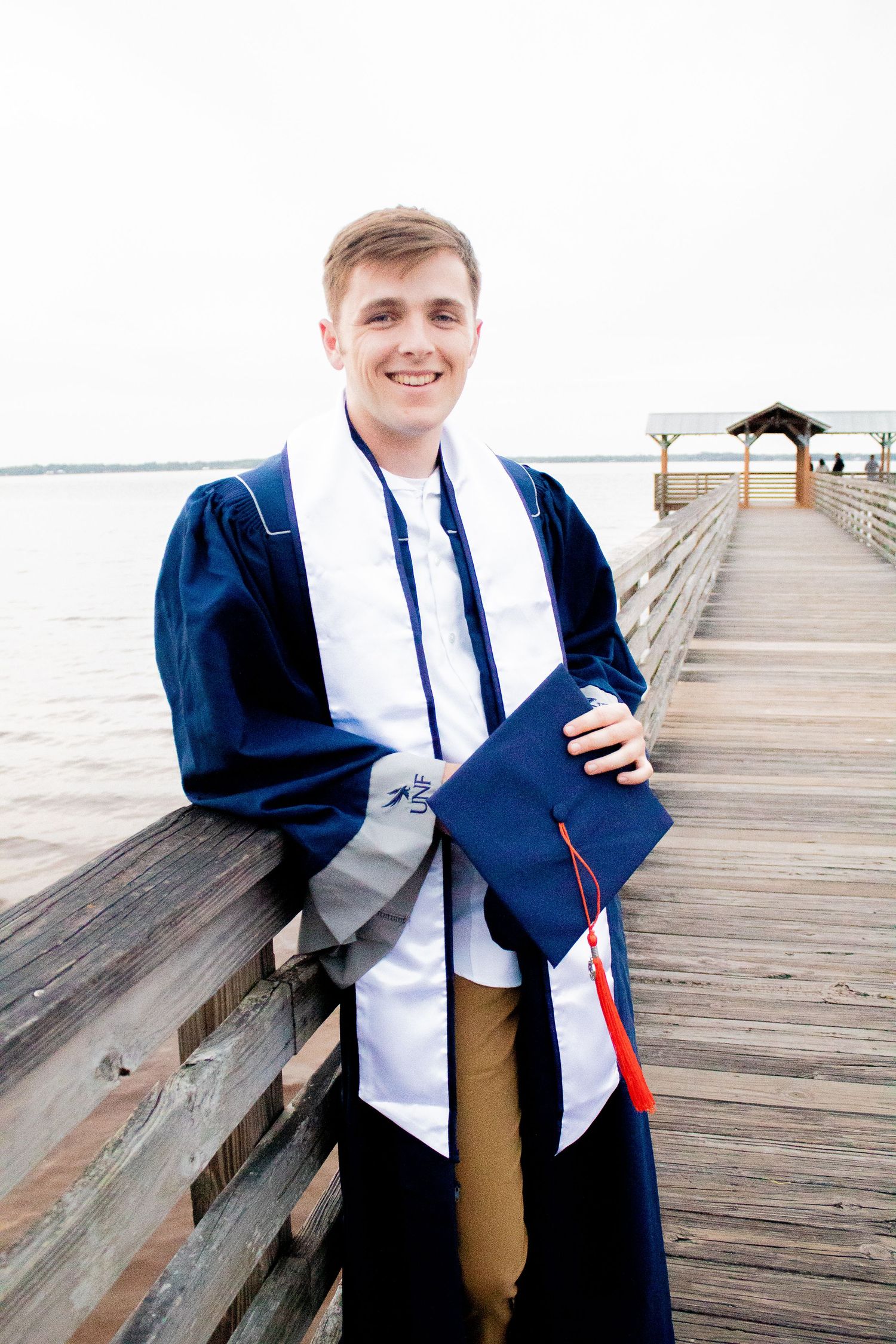 Graduation photo of a guy leaning back on a bridge, holding his cap while wearing his gown.