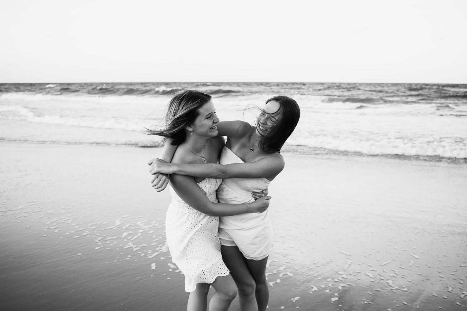 Black-and-white photo of best friends laughing and embracing each other while standing on the beach, with waves and sand in the background.