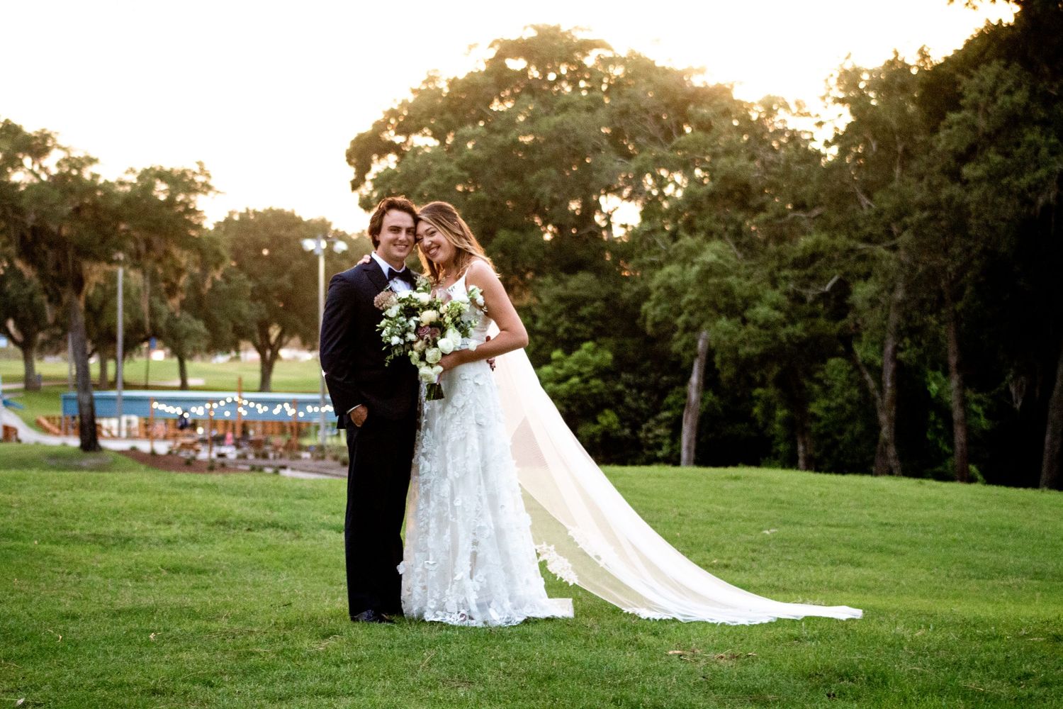 Photo of the bride leaning on the groom in a grassy field, with a stunning sunset backdrop.