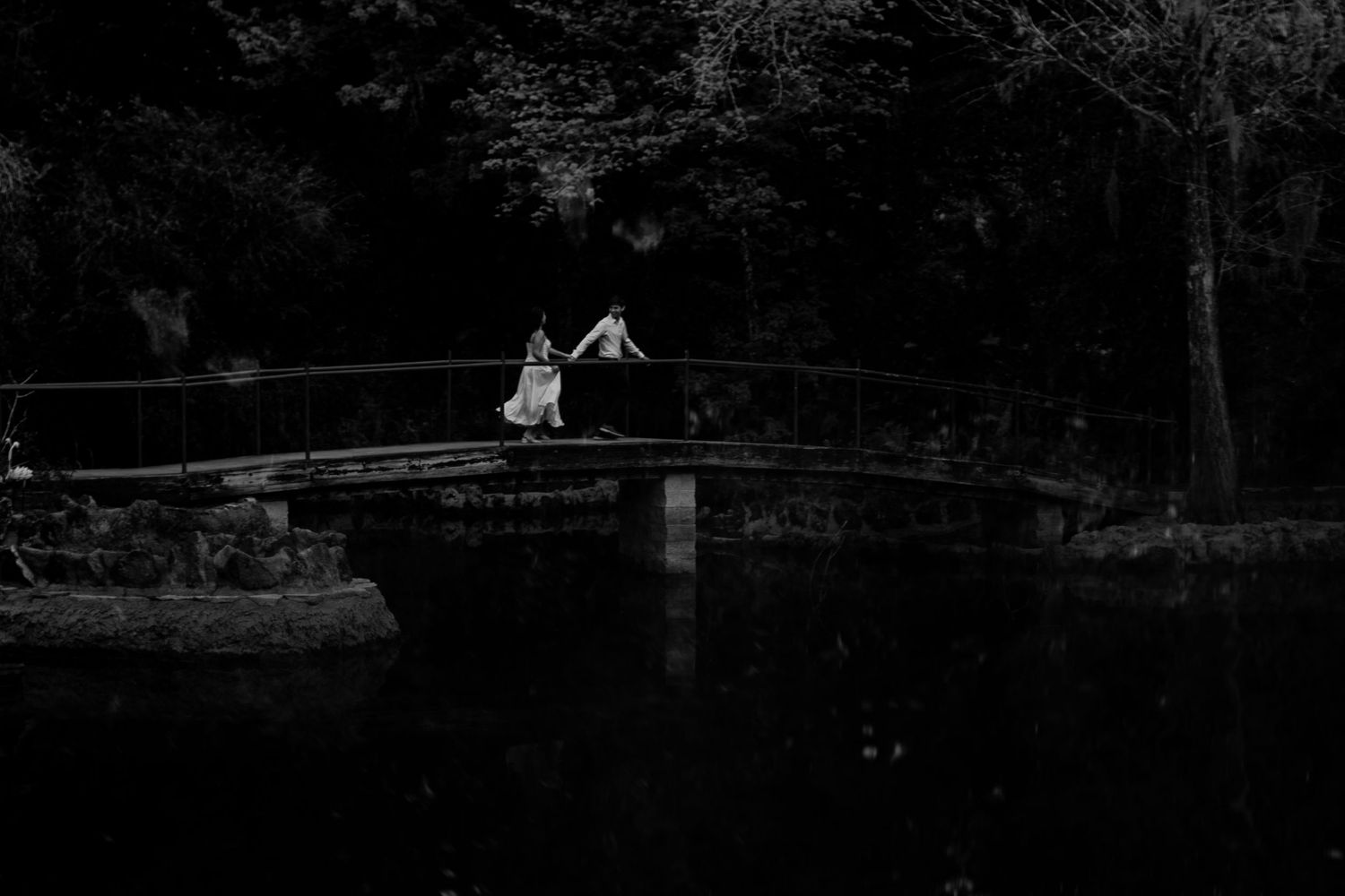 Two lovers run hand in hand on a bridge—she glances back, wild and free, like they’re running toward forever. B&W magic.