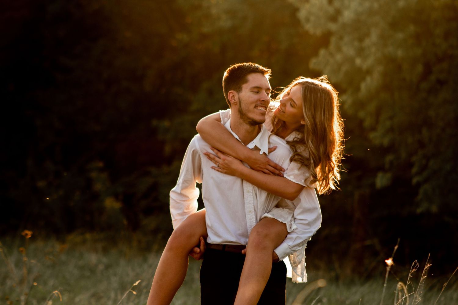 Engagement photo of a woman piggybacking on her fiancé, smiling at him as he looks back at her with a sunset-lit field in the background.