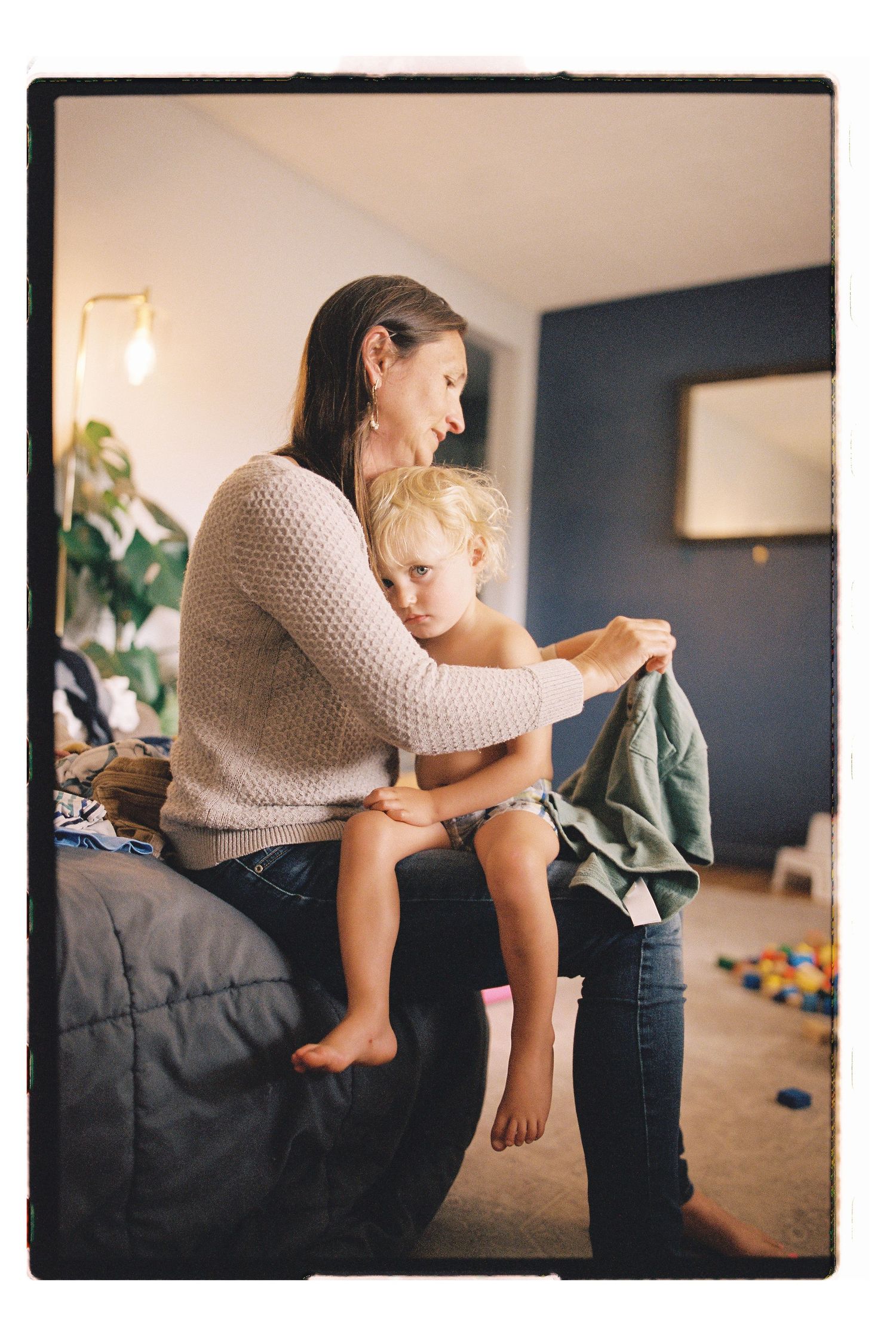 A mother helps her young blonde toddler get dressed while sitting on a bed in a cozy home bedroom.