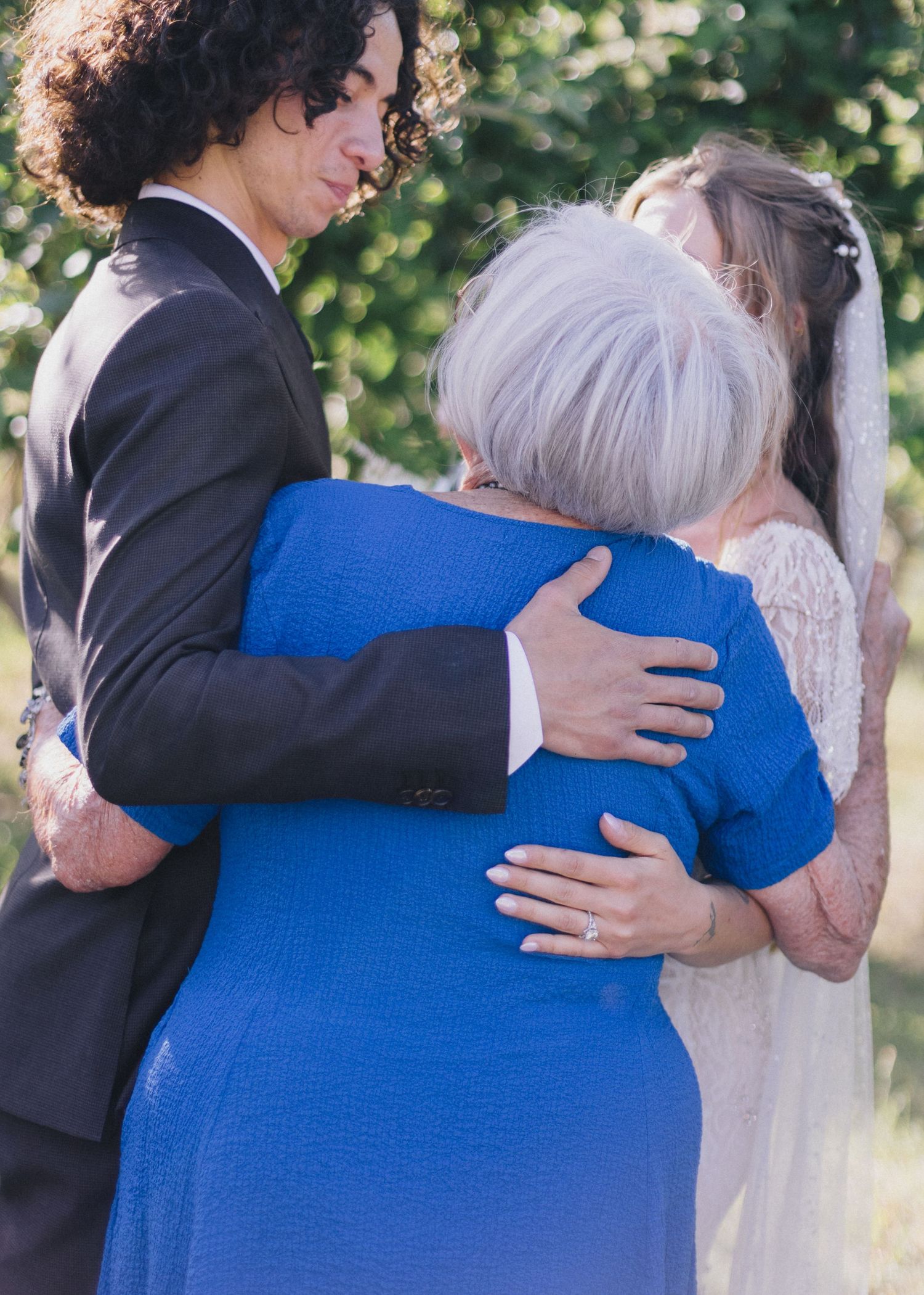 Groom in black suit hugging elderly woman in blue dress at outdoor wedding ceremony with bride in lace gown nearby.