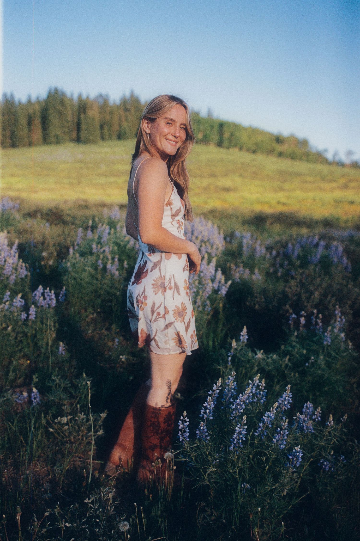 Young woman in floral dress and brown cowboy boots smiling in a purple wildflower meadow with pine trees.