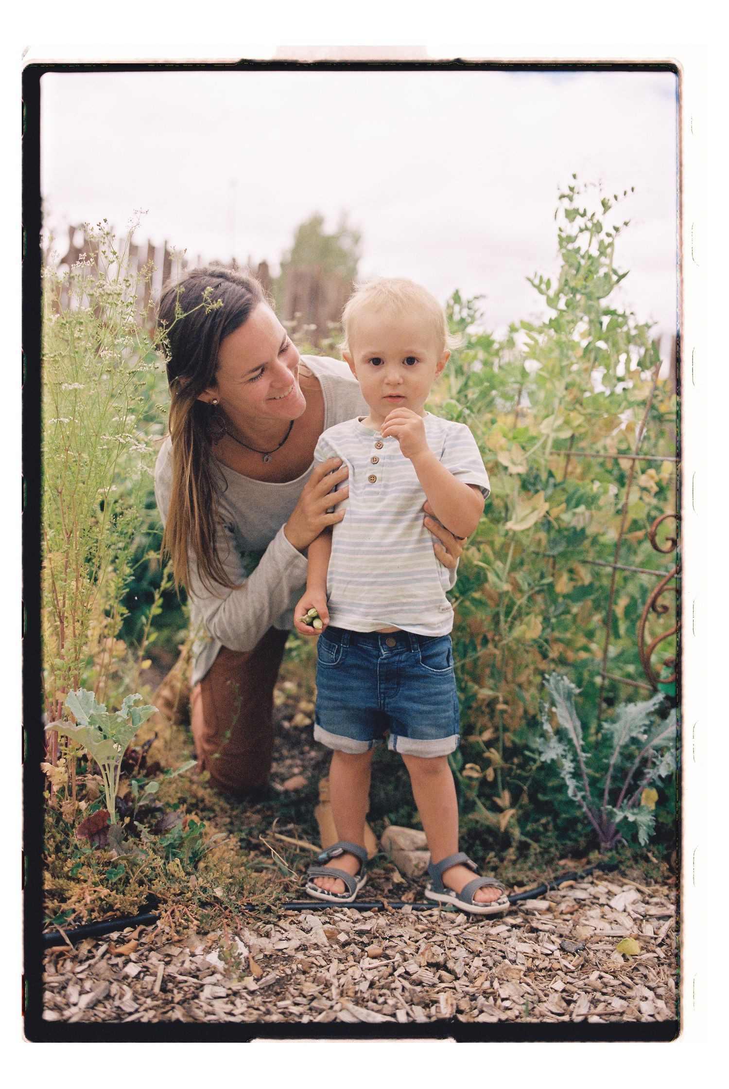 A mother and toddler boy explore a lush garden together, with the child eating fresh produce among green plants.