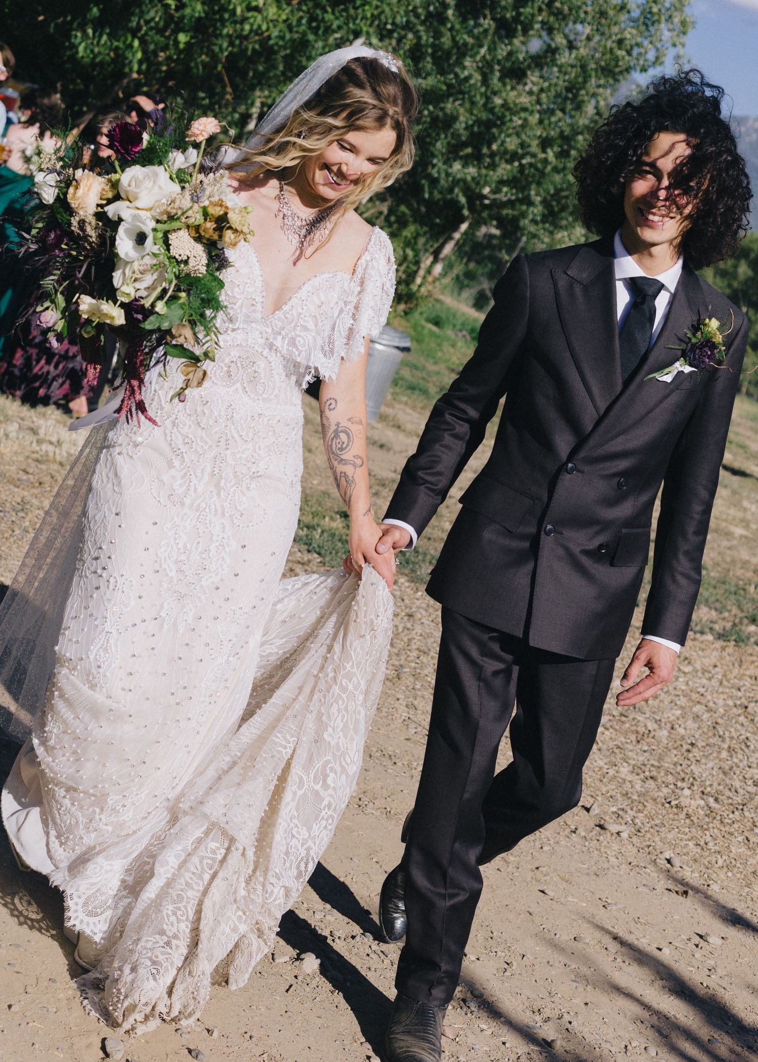 A bride in lace gown with veil holds bouquet while walking hand-in-hand with groom in black suit outdoors.