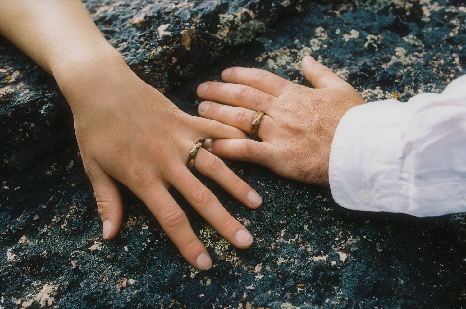 Two hands with gold wedding rings touching on a rocky surface, symbolizing love and commitment.