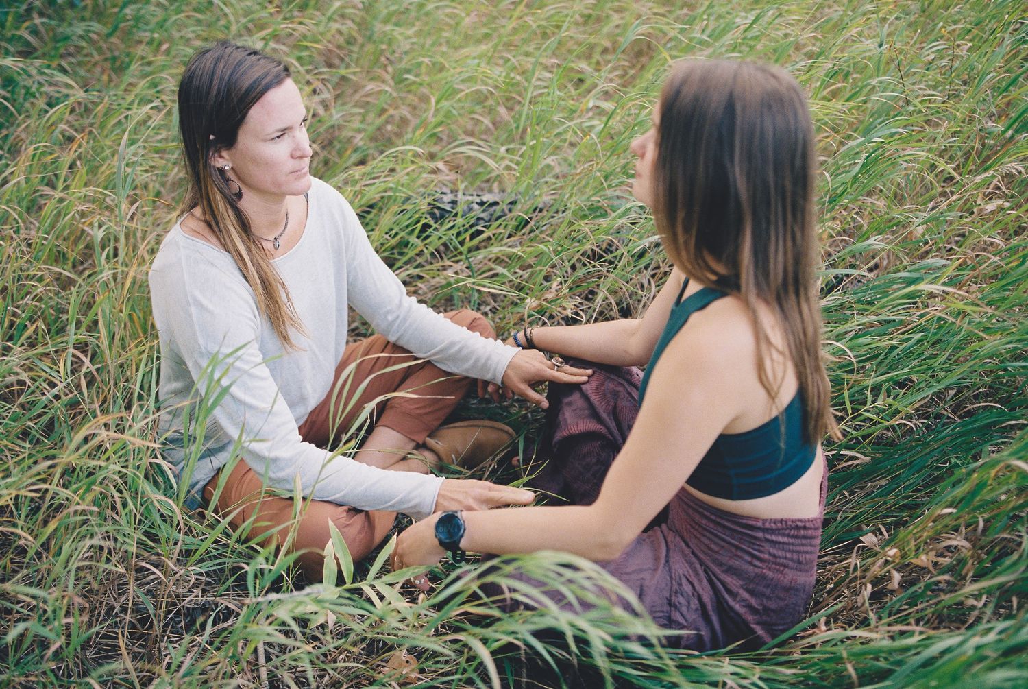 Two women sitting in tall grass, facing each other and holding hands in a peaceful outdoor setting.