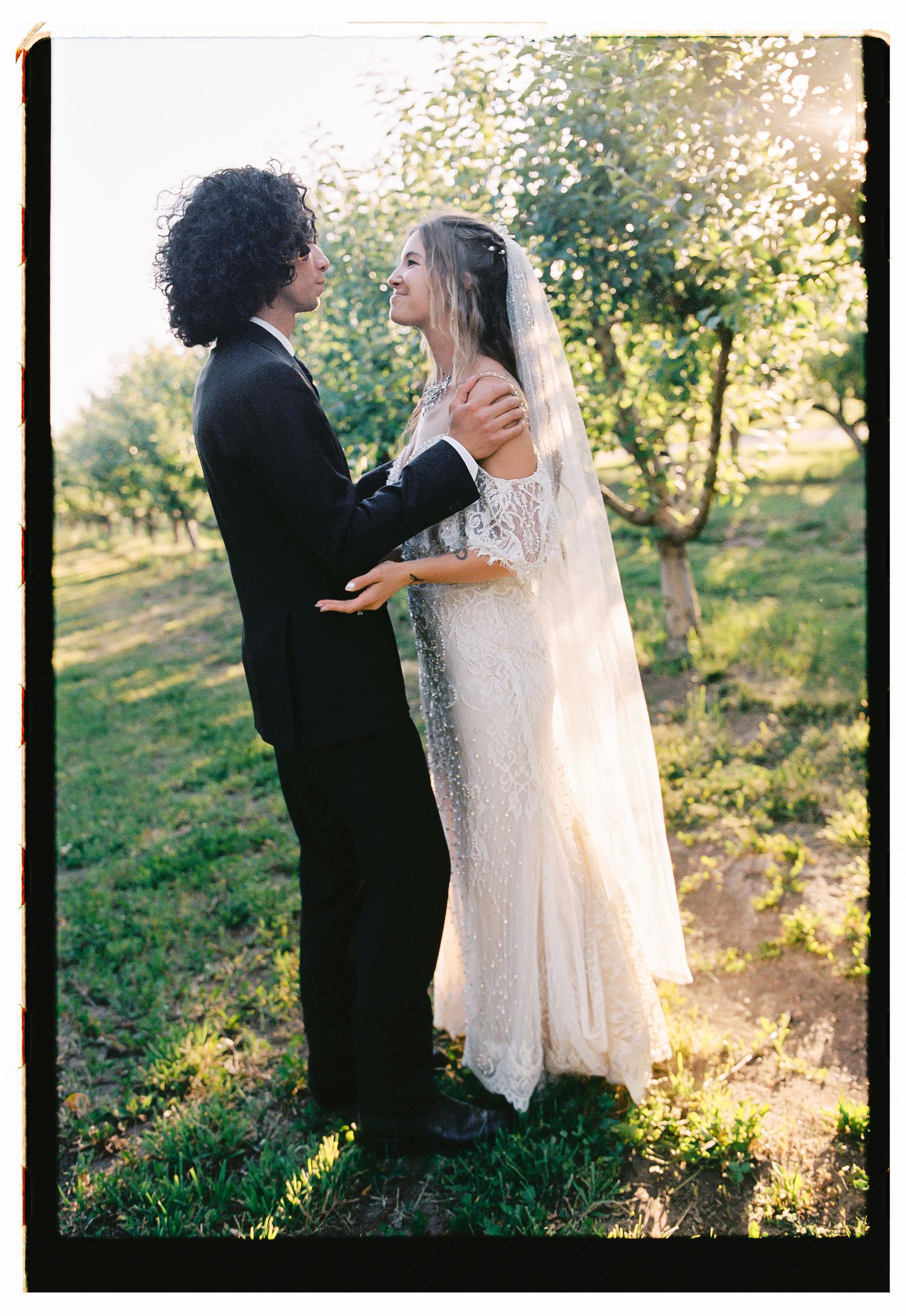 A bride in a lace gown and veil shares a romantic moment with her groom in a black suit among sunlit orchard trees.