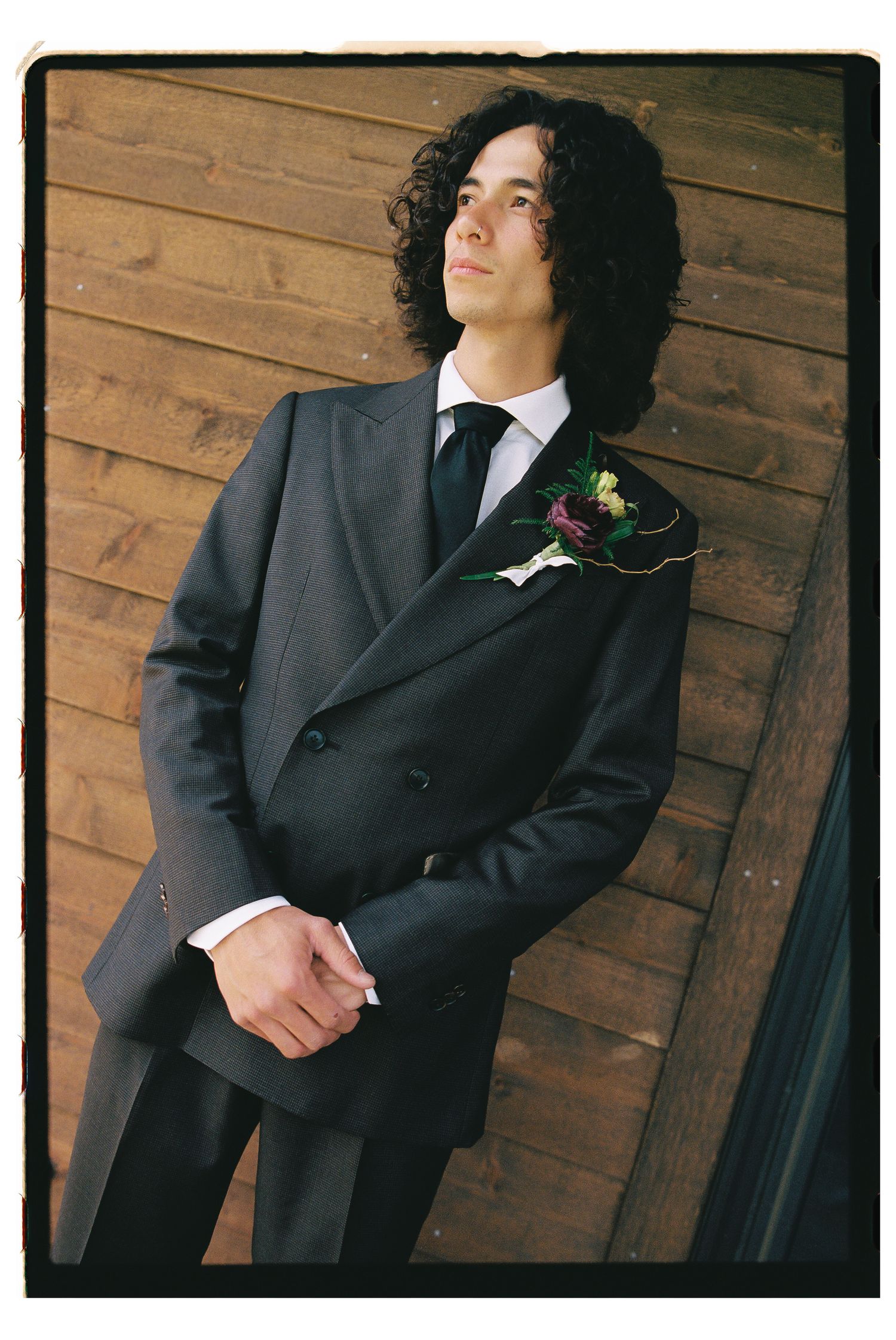 Young man in dark double-breasted suit with boutonniere, posing against wooden wall with curly hair.