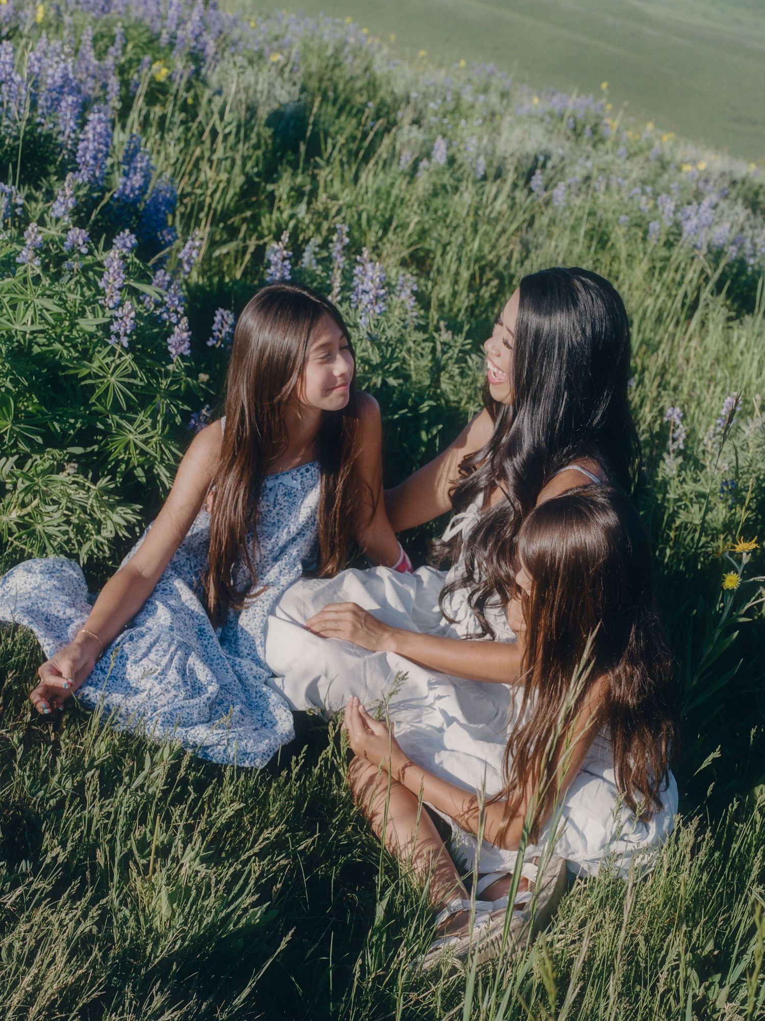 A woman and two girls in floral dresses sit together in a lush green field with purple wildflowers.