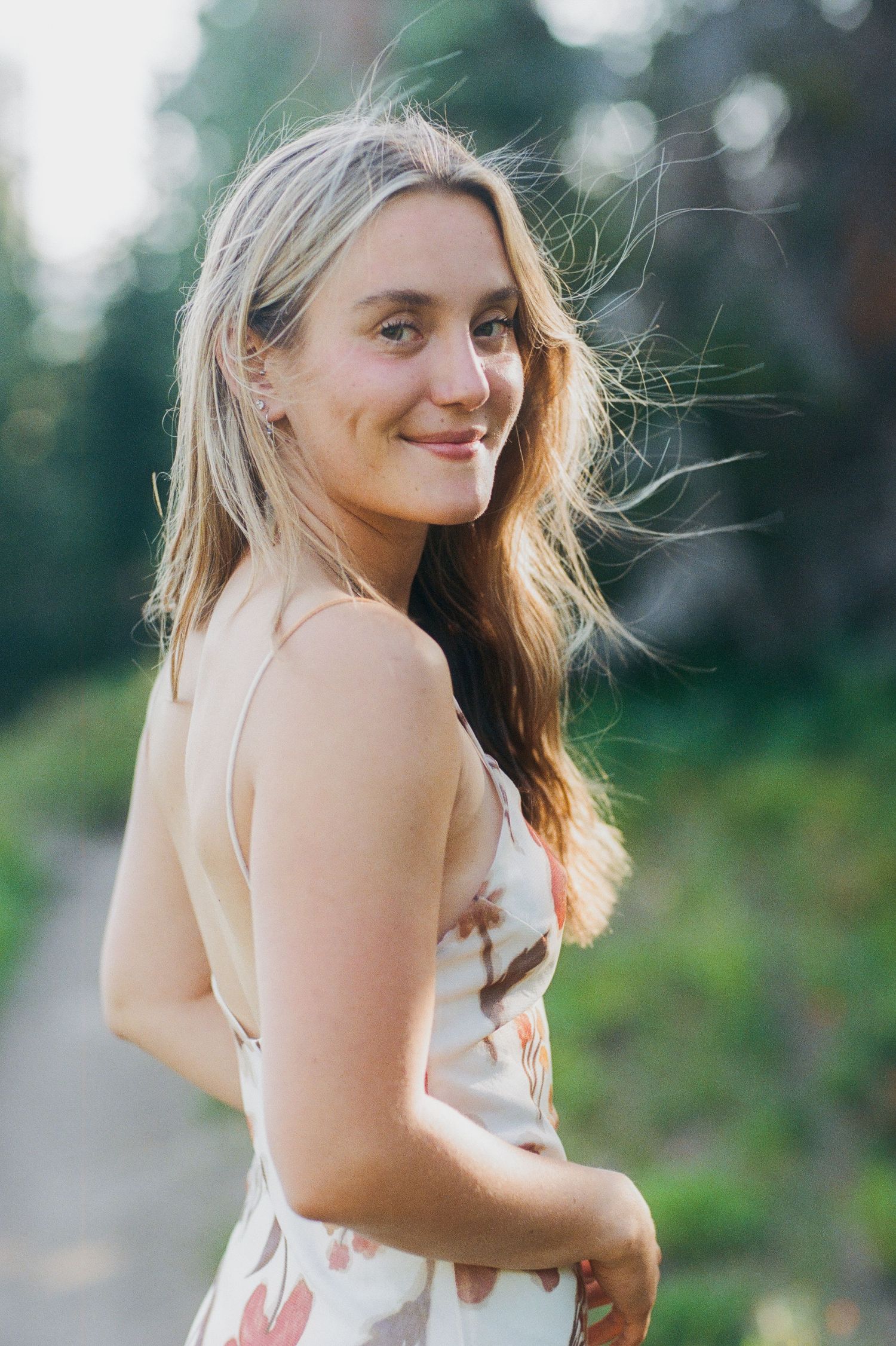 Young woman in floral dress smiling outdoors with long blonde hair blowing in the wind on a nature path.