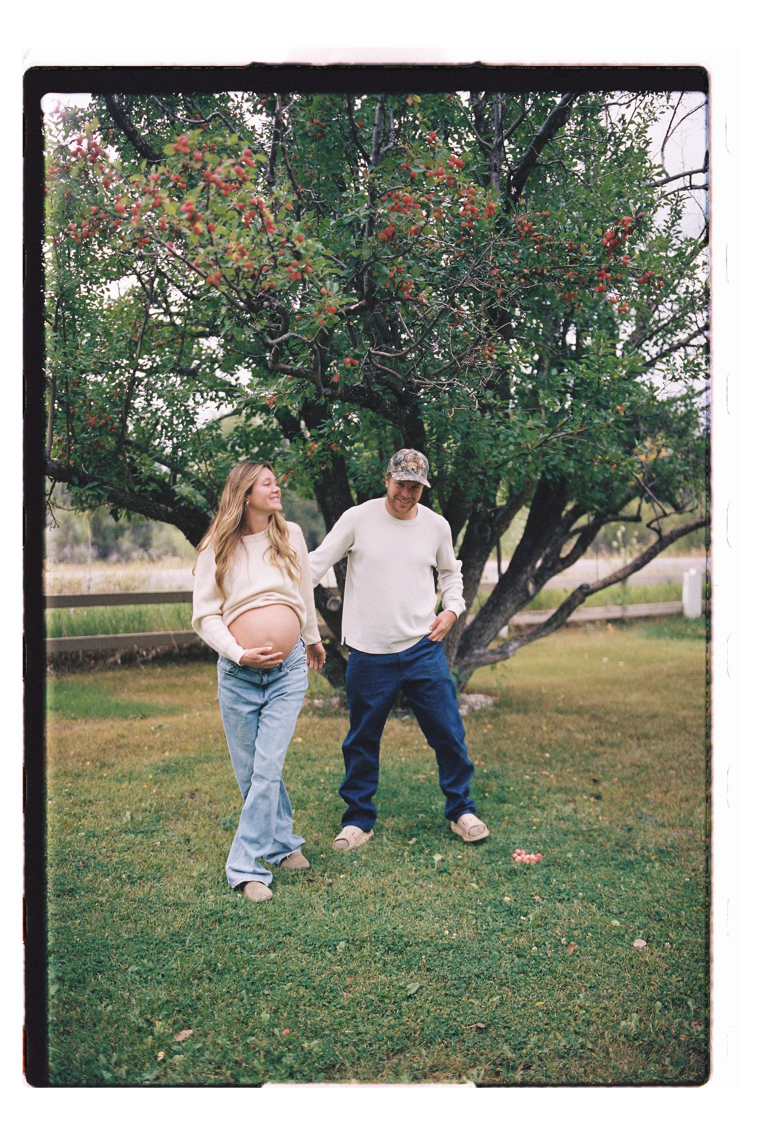 Pregnant woman and man standing together outdoors under a tree on green grass during a maternity photo shoot.