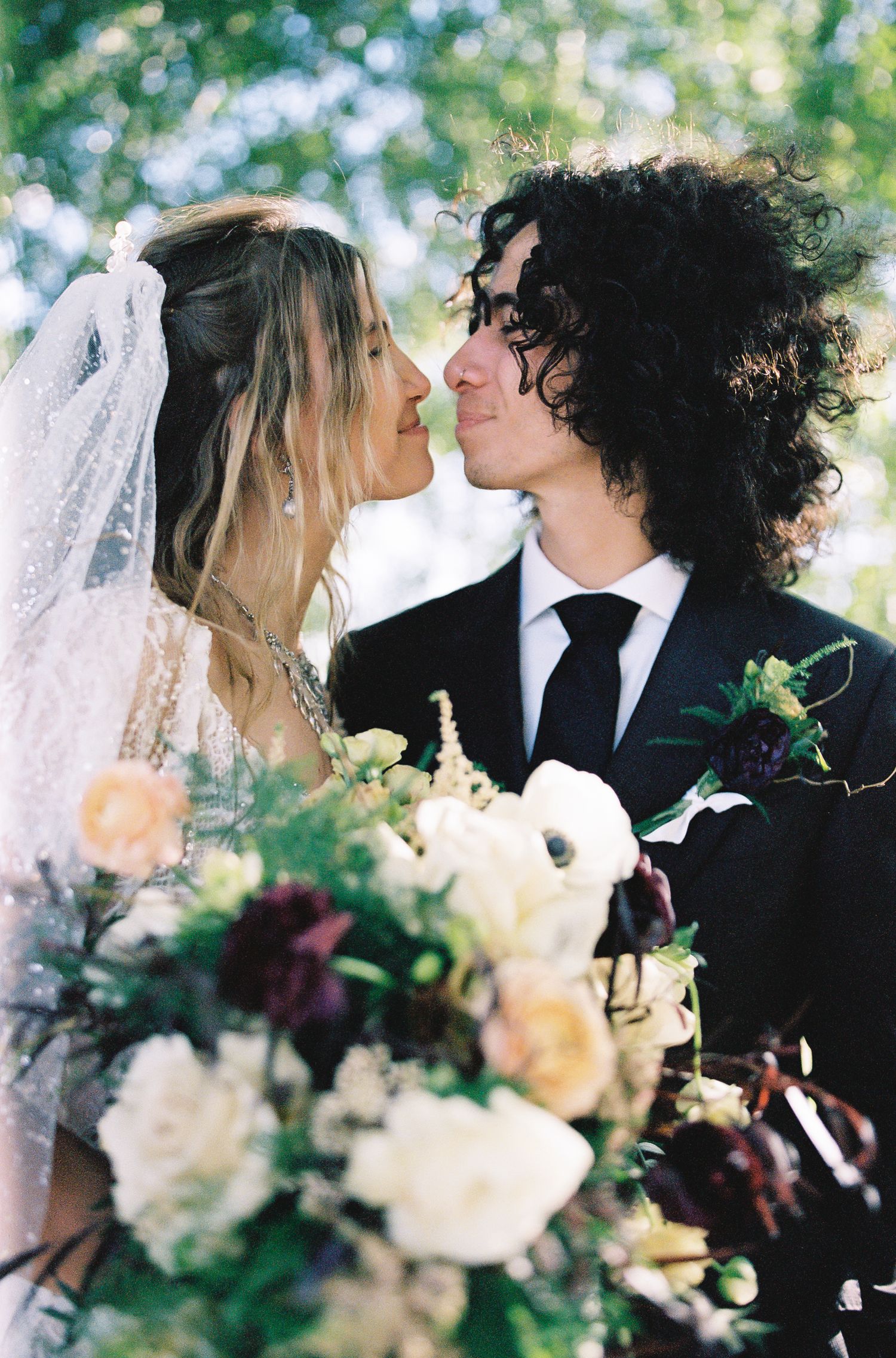 A bride and groom share a romantic moment, nearly kissing, with a lush floral bouquet in the foreground.