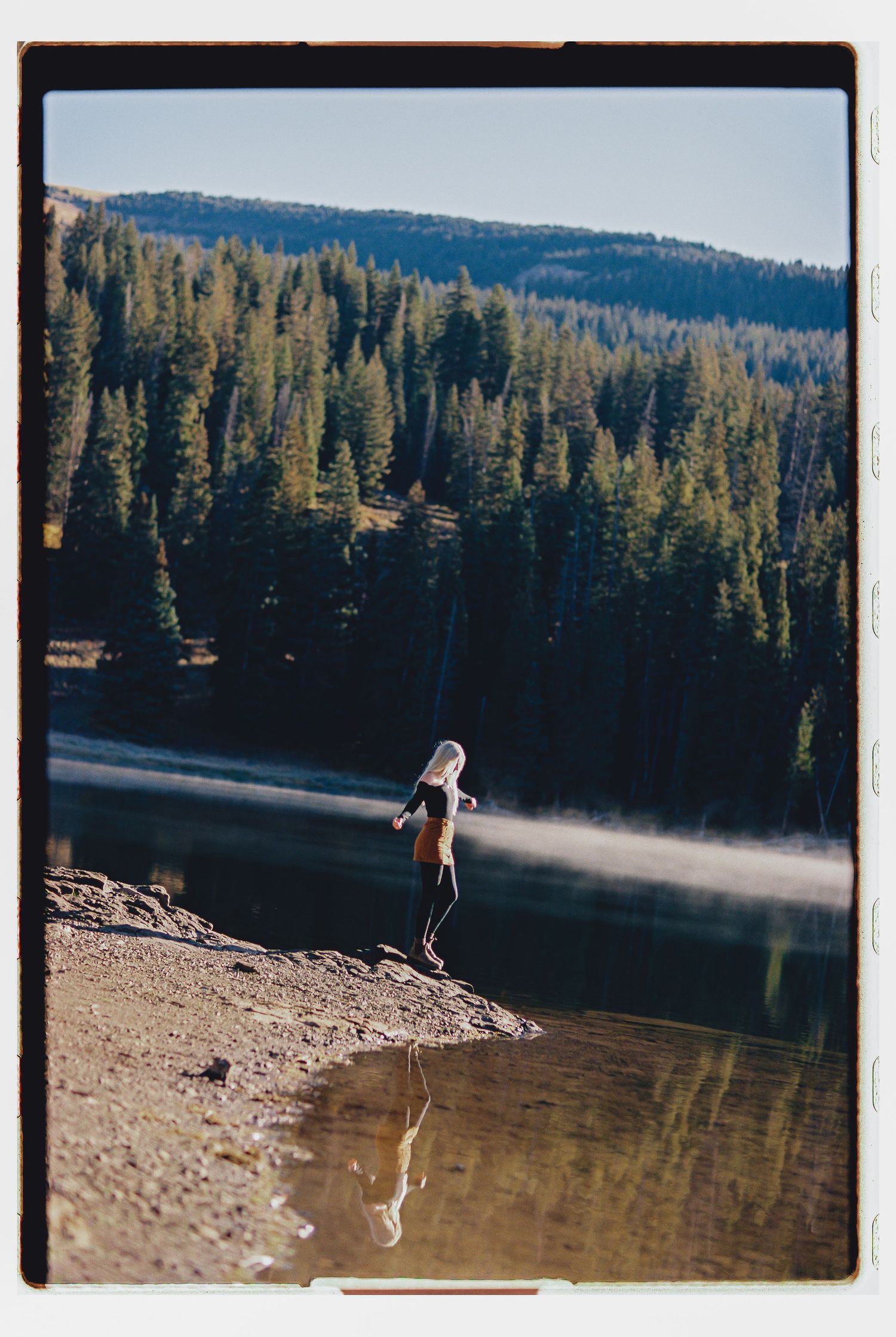 Person standing at the edge of a mountain lake surrounded by pine trees, with a clear reflection in the calm water.