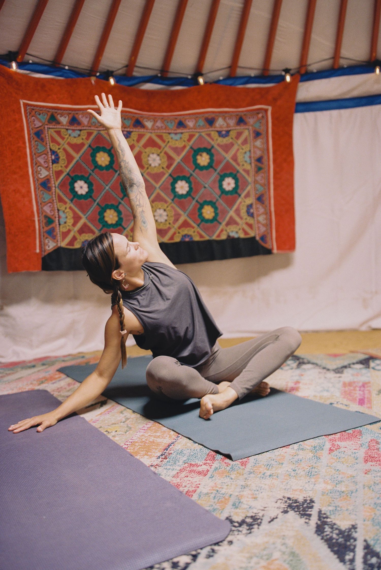 Woman practicing yoga in a yurt, performing a seated side stretch with one arm raised, on a blue mat with colorful tapestry backdrop.