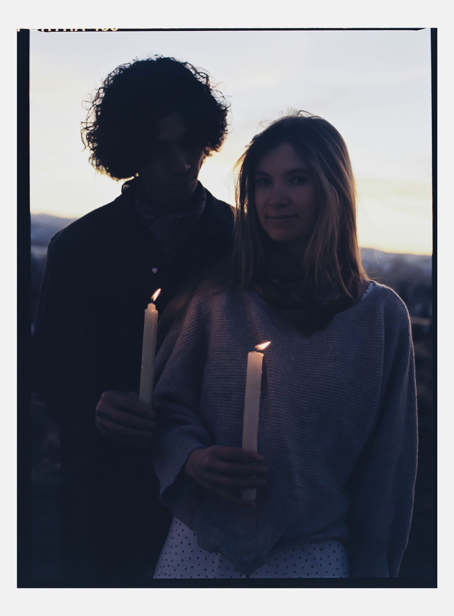 Couple holding lit candles at dusk in a romantic outdoor setting, soft twilight glow illuminating their faces.