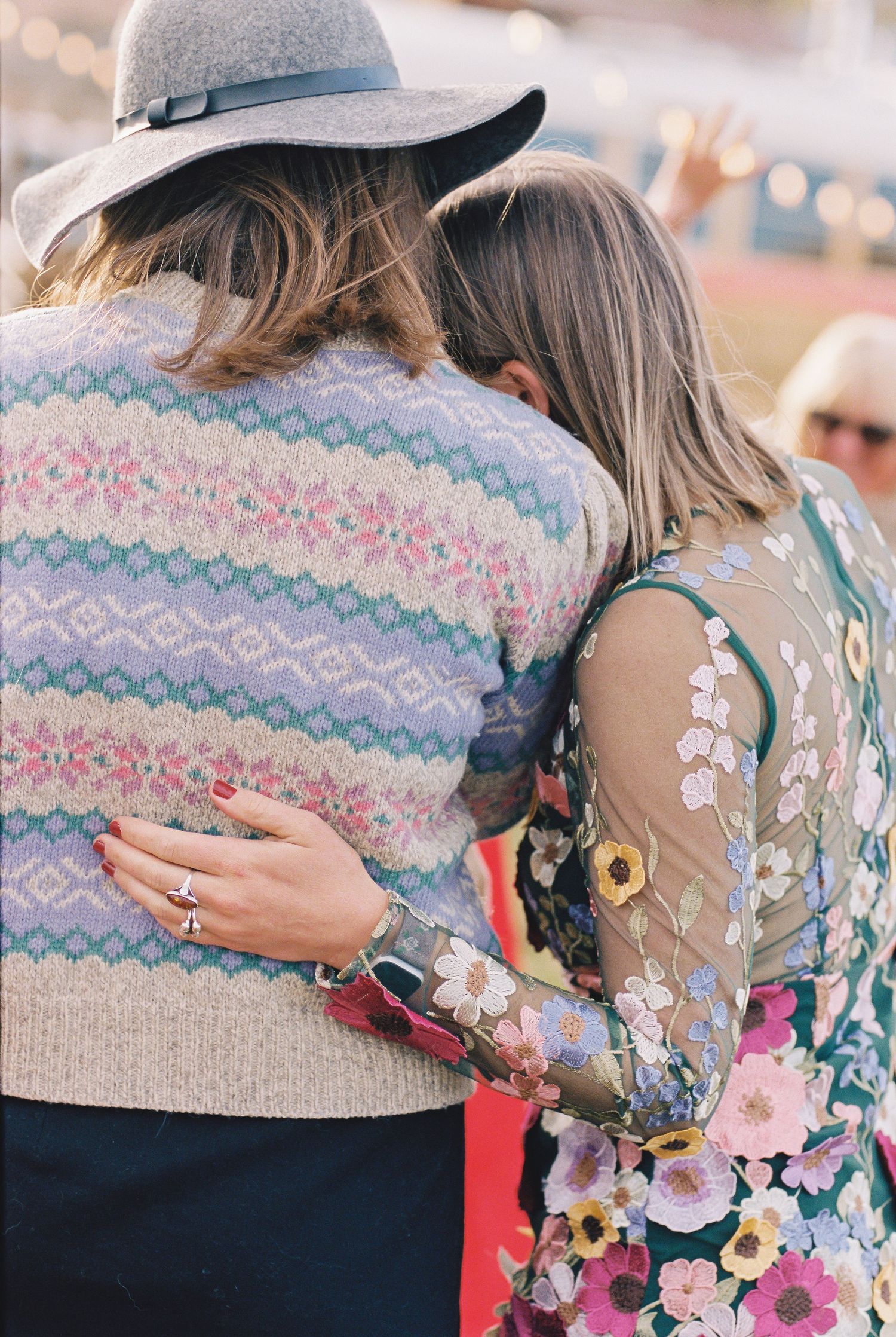 Two women embracing warmly, one wearing a floral Fair Isle sweater and hat, the other in a floral embroidered dress.