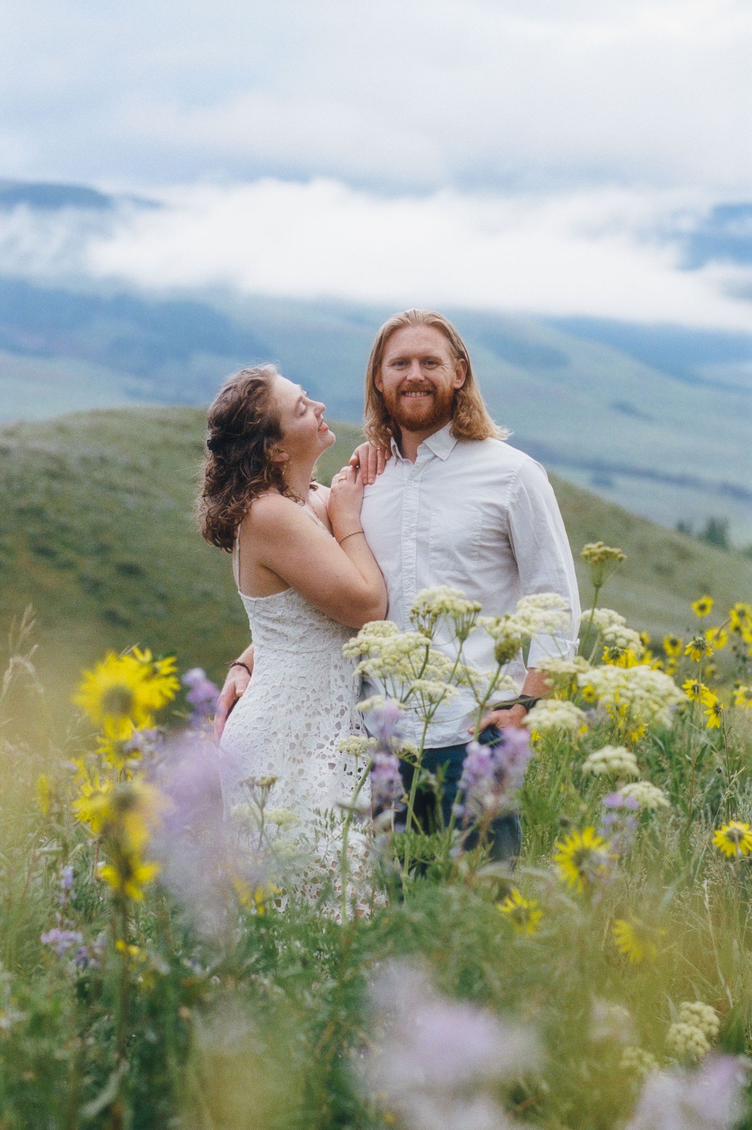 Couple embracing in a wildflower meadow with mountain views, woman in white dress, man with red hair and beard.