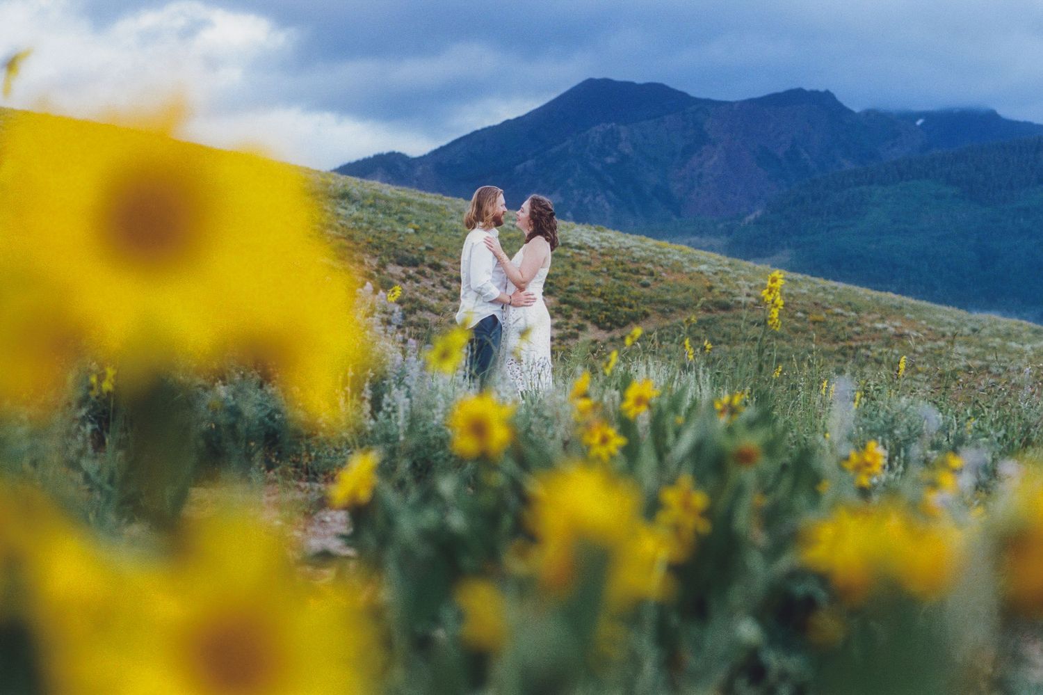 Couple embraces in a field of yellow wildflowers with mountain backdrop on a cloudy day.