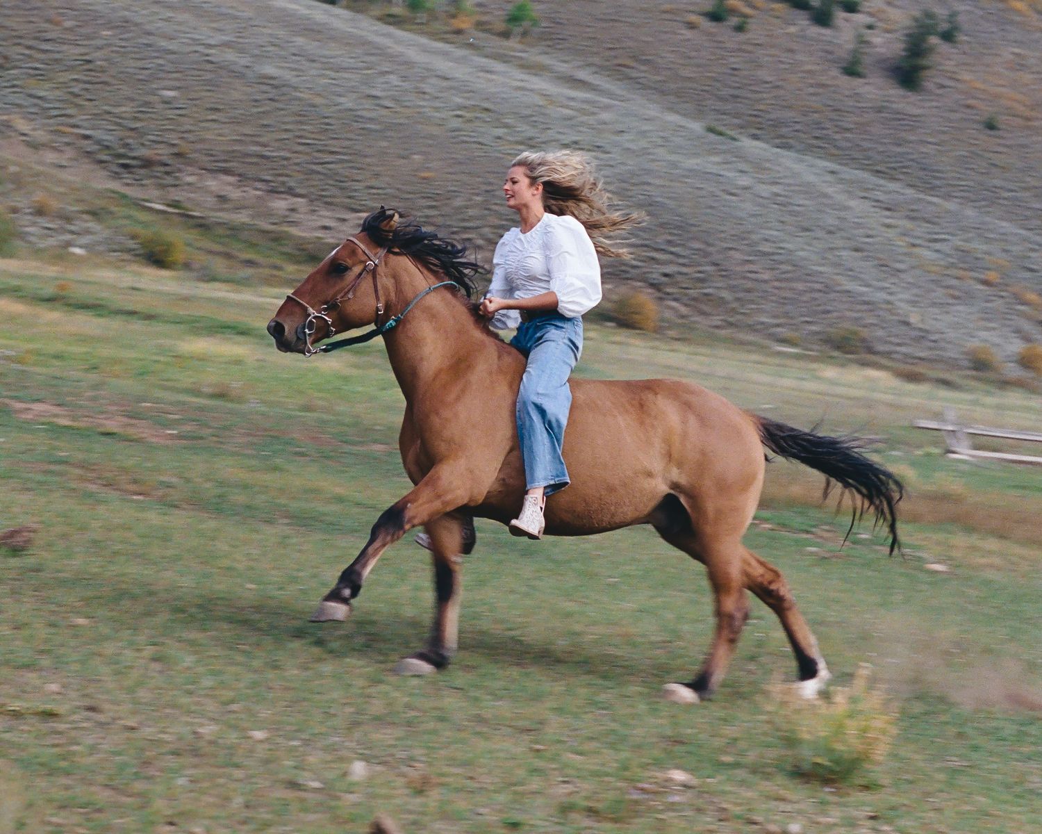 Woman with flowing hair riding a brown horse bareback across a green mountain valley field.