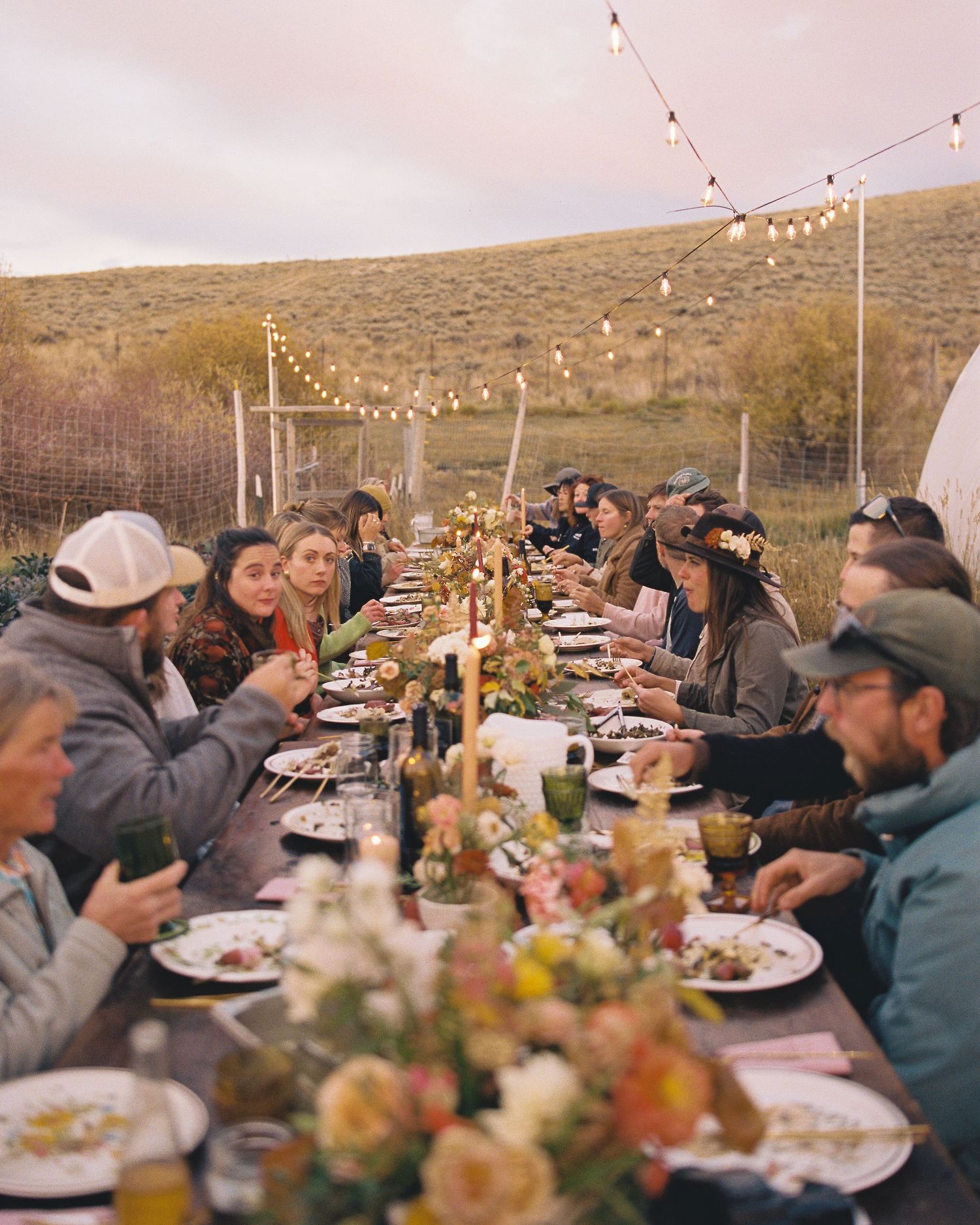 Guests dining at a long outdoor table with string lights at a rustic farm dinner party at dusk.