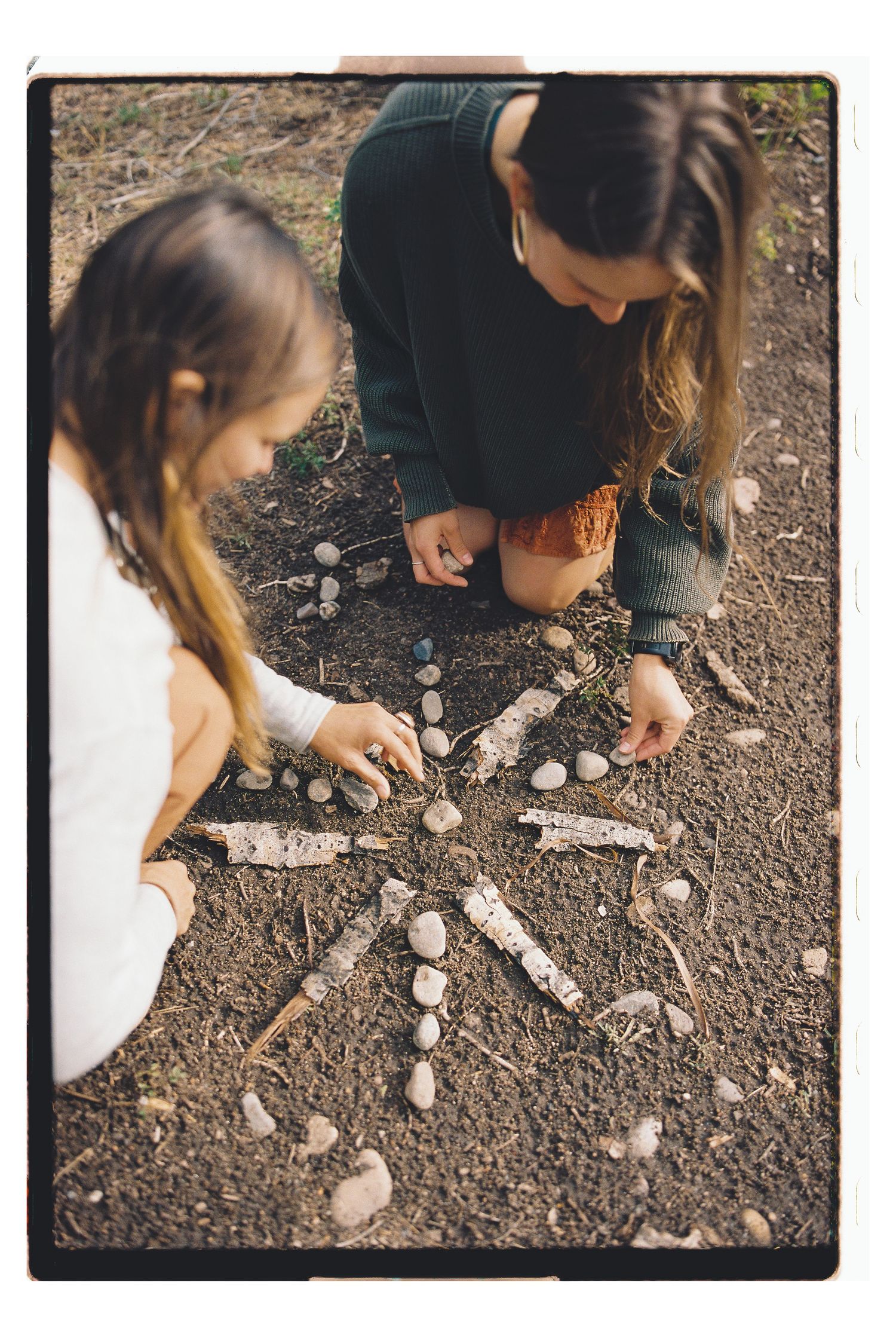 Two girls arranging sticks and stones into a mandala pattern on the ground outdoors.