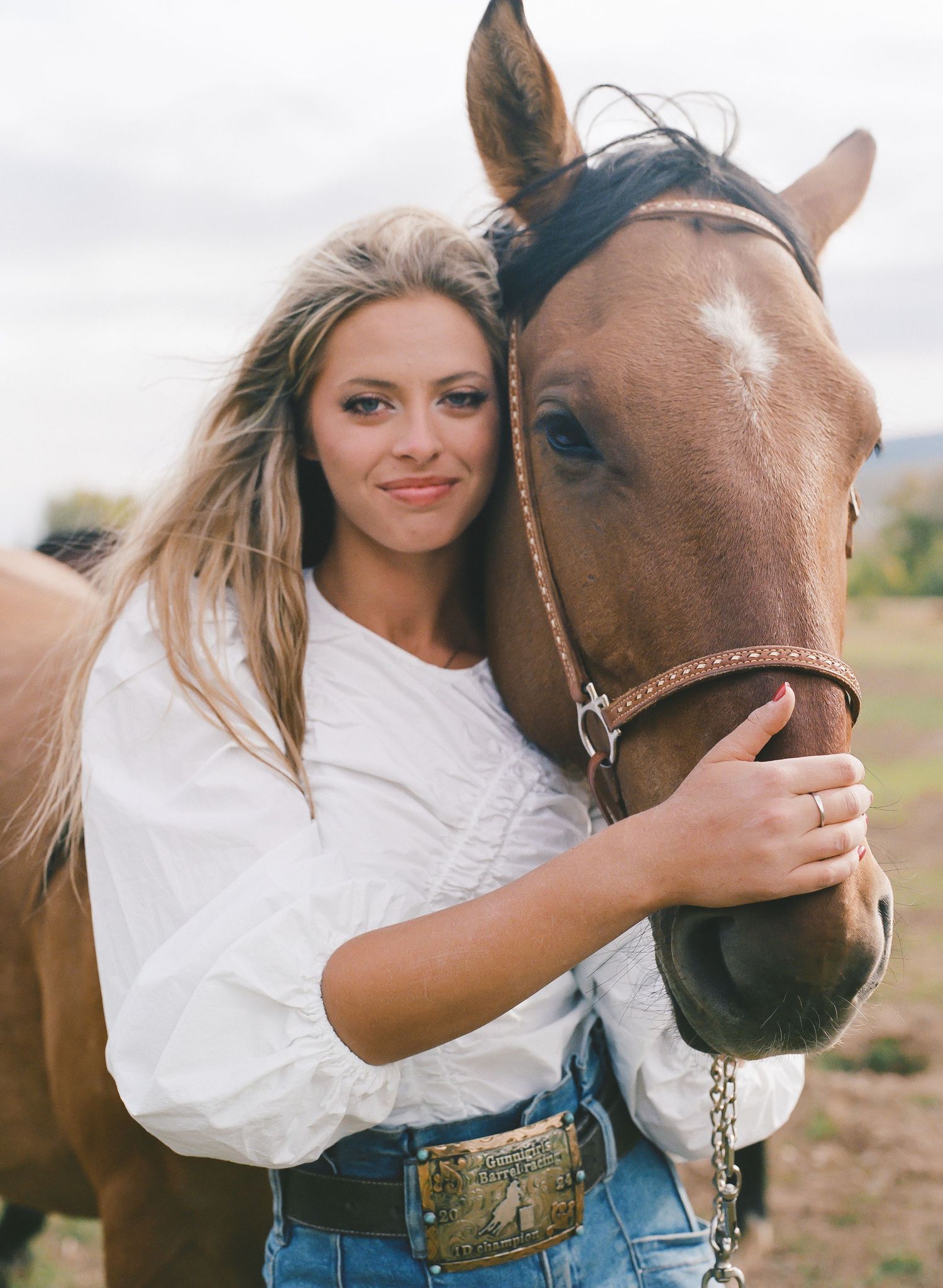 Young woman in white top and jeans hugging a brown horse in a field, wearing a decorative belt.