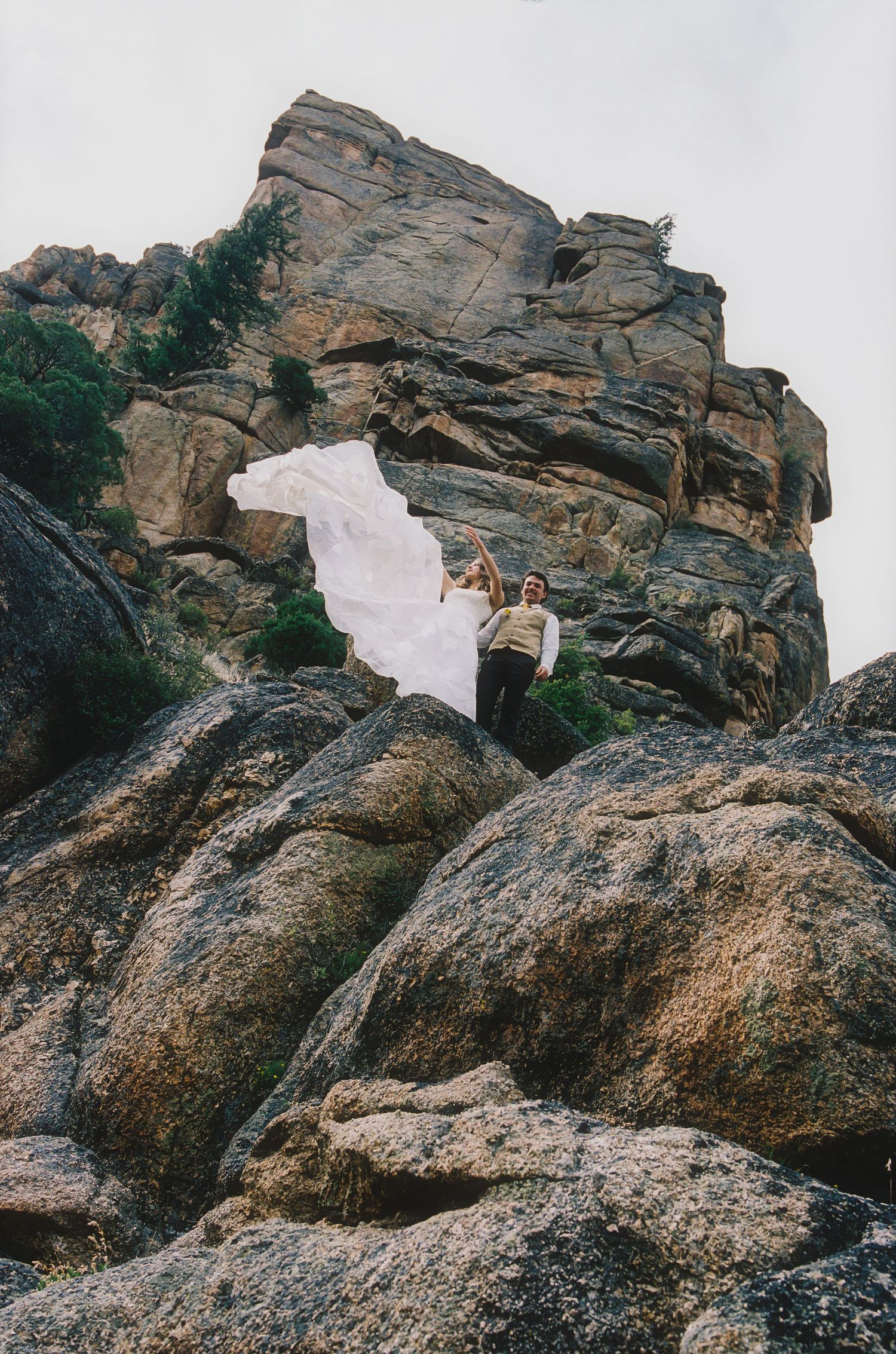 Couple in wedding attire standing on rocky cliffs, bride's white dress flowing dramatically in the wind against rugged stone formations.