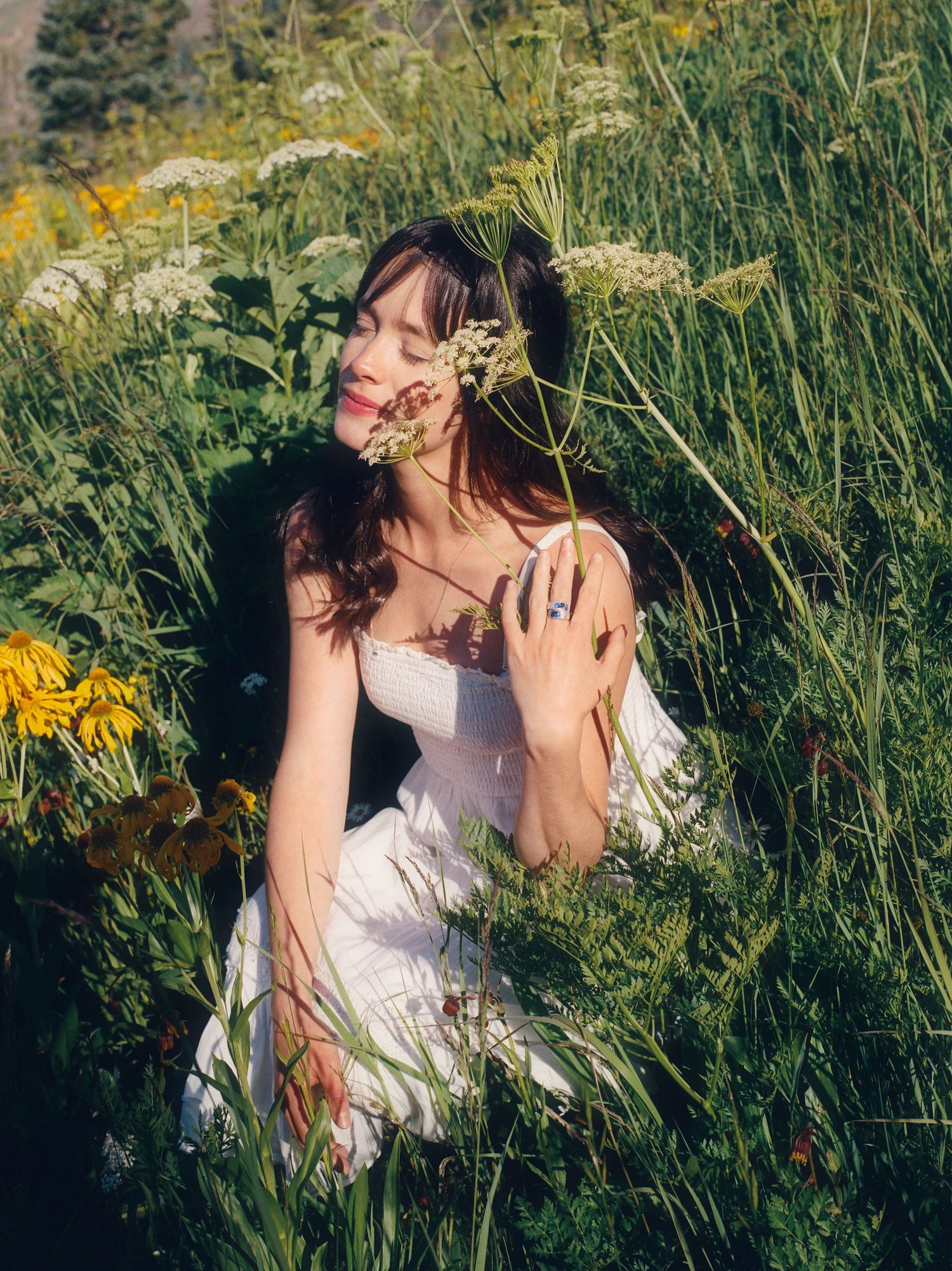 Woman in white dress sitting among wildflowers and tall green grass, holding blooms close to her face.