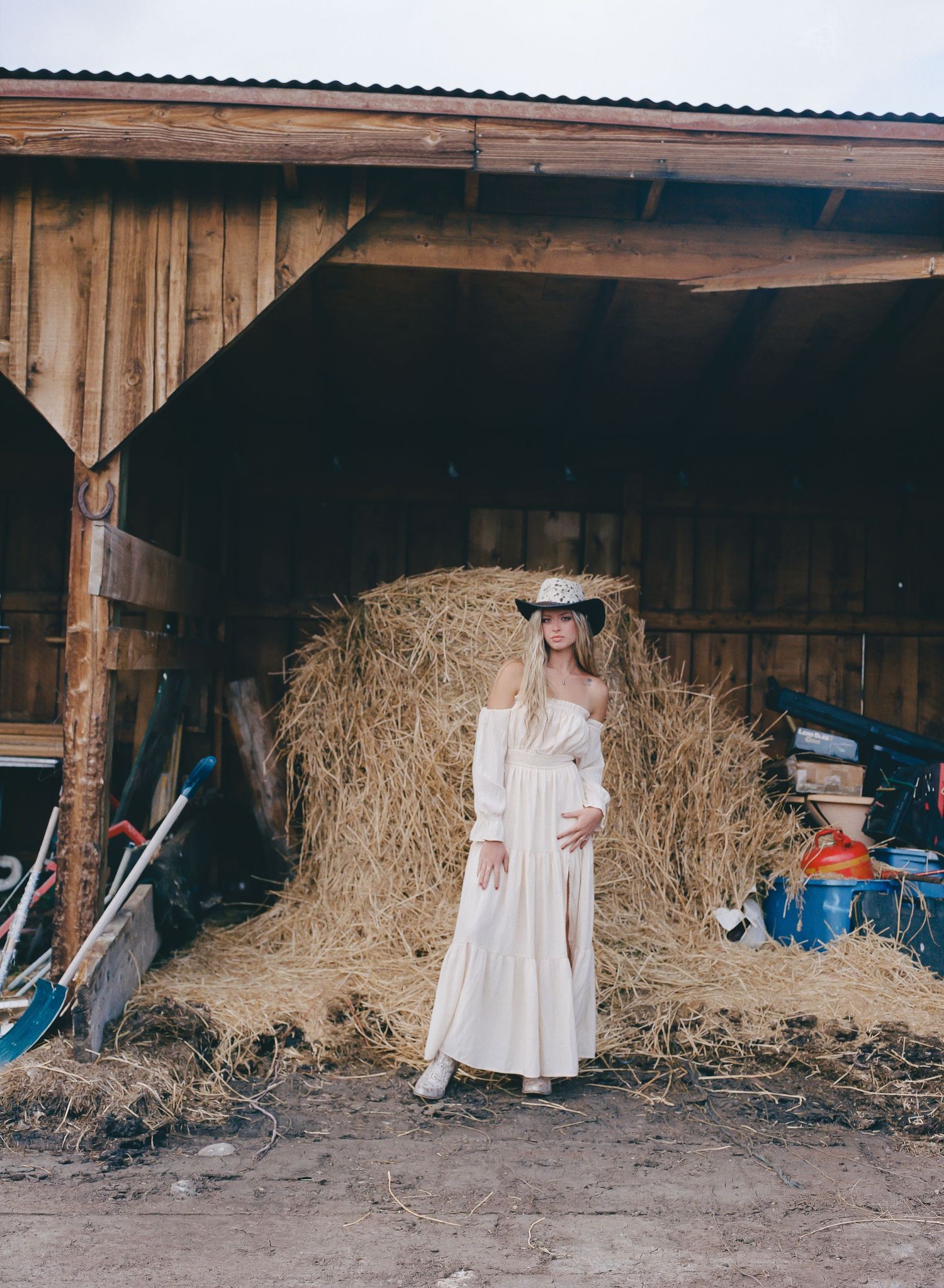 Woman in white dress and cowboy hat poses in front of hay bales inside a rustic wooden barn.