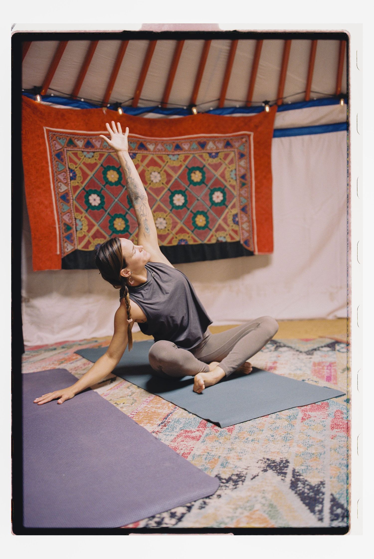 Woman practicing yoga in a decorated yurt with colorful tapestries, performing a seated side stretch on a gray mat.