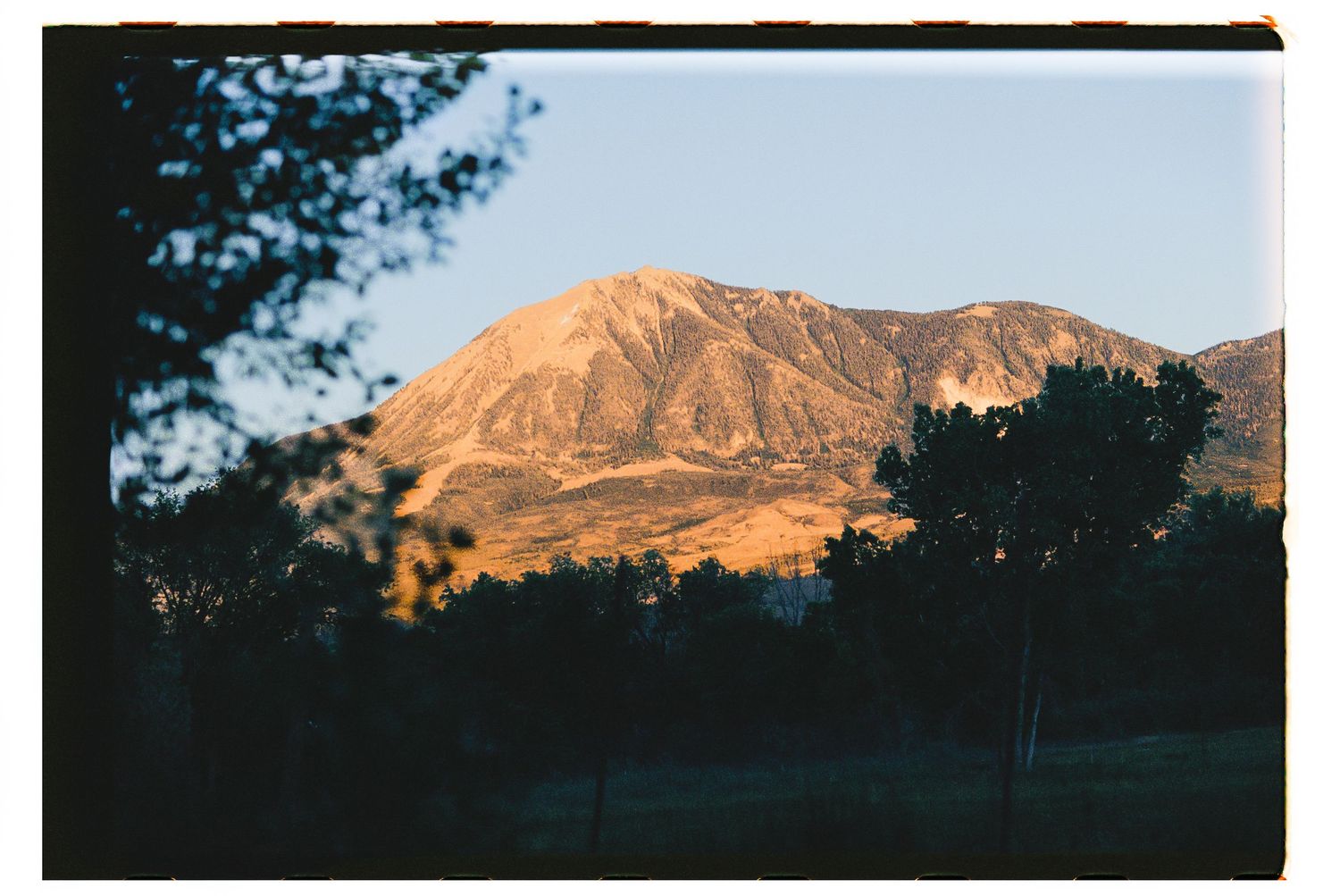 Sunlit mountain peak glowing in warm golden light at dusk, framed by silhouetted trees in the foreground.