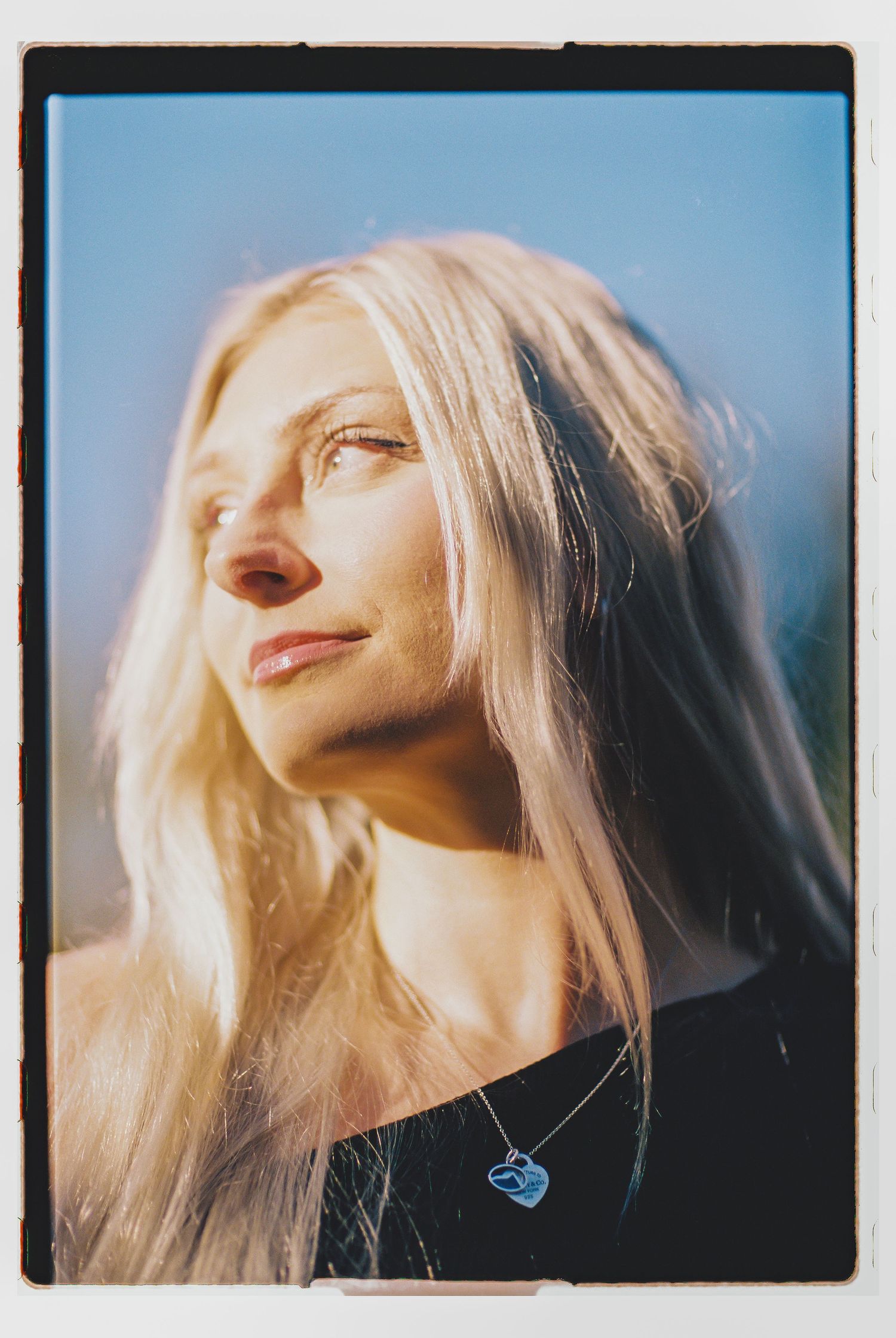 Blonde woman with long hair wearing a blue necklace, bathed in warm sunlight against a clear blue sky.
