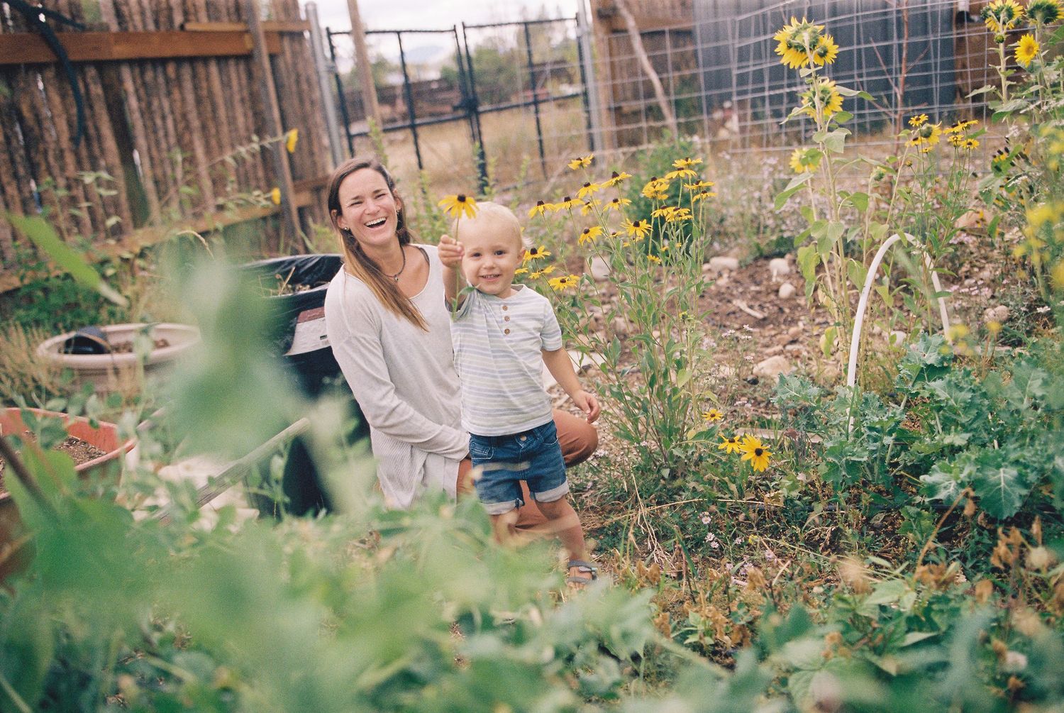 A laughing woman and toddler stand together in a lush garden with yellow wildflowers and green plants.