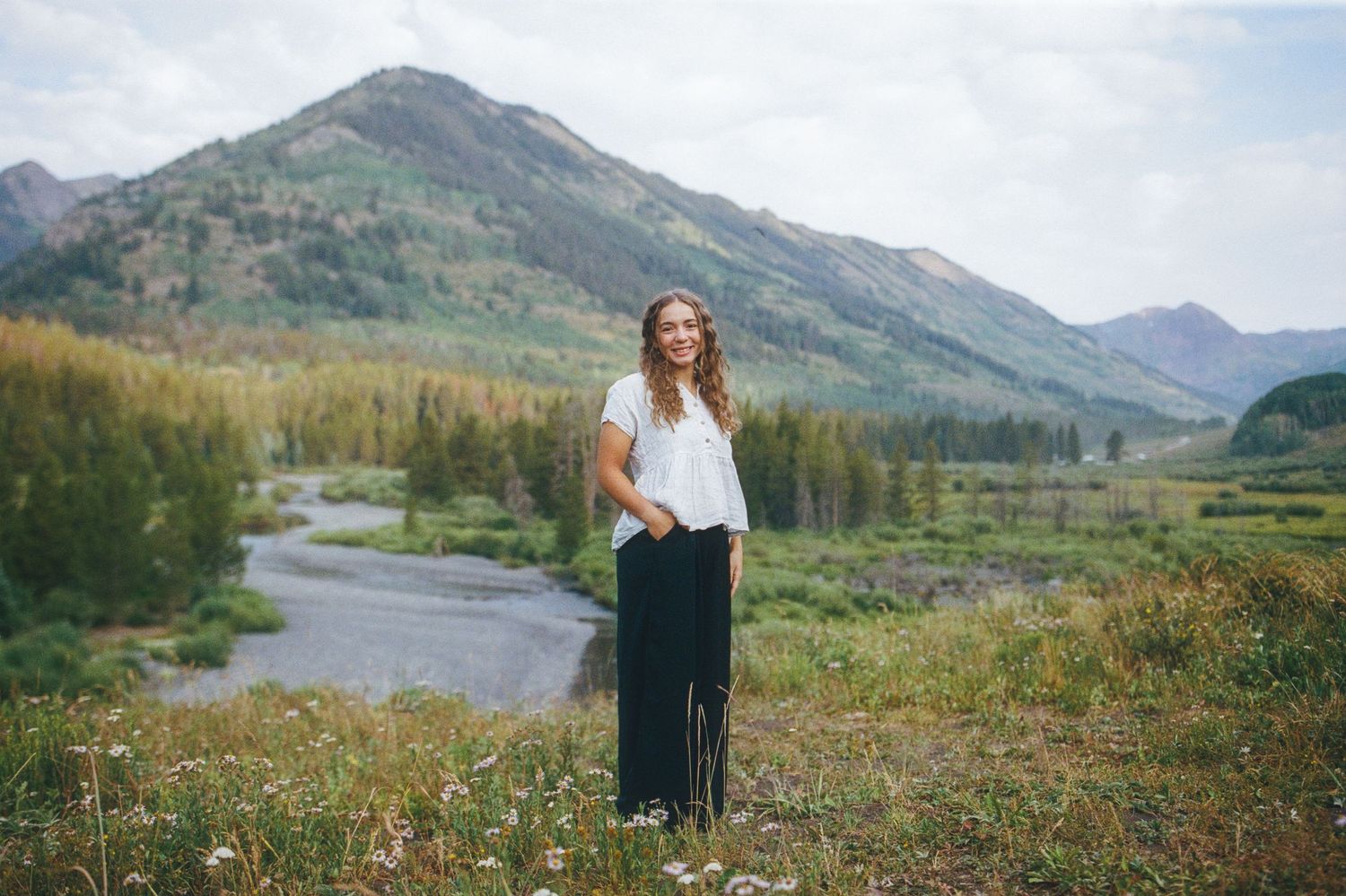 Woman smiling in white top and black pants standing in a scenic mountain meadow with a stream and trees.