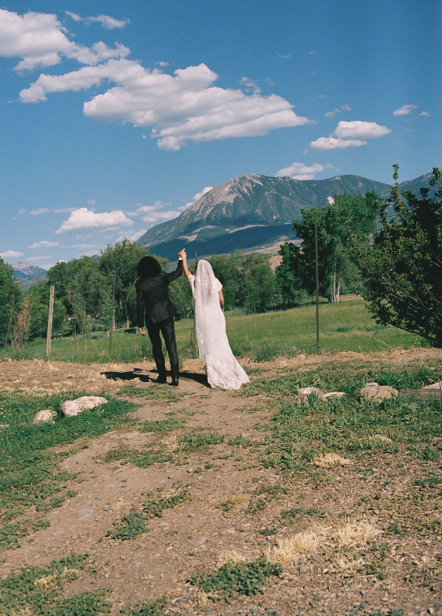 A bride and groom hold hands walking toward a stunning mountain landscape under a blue sky on their wedding day.