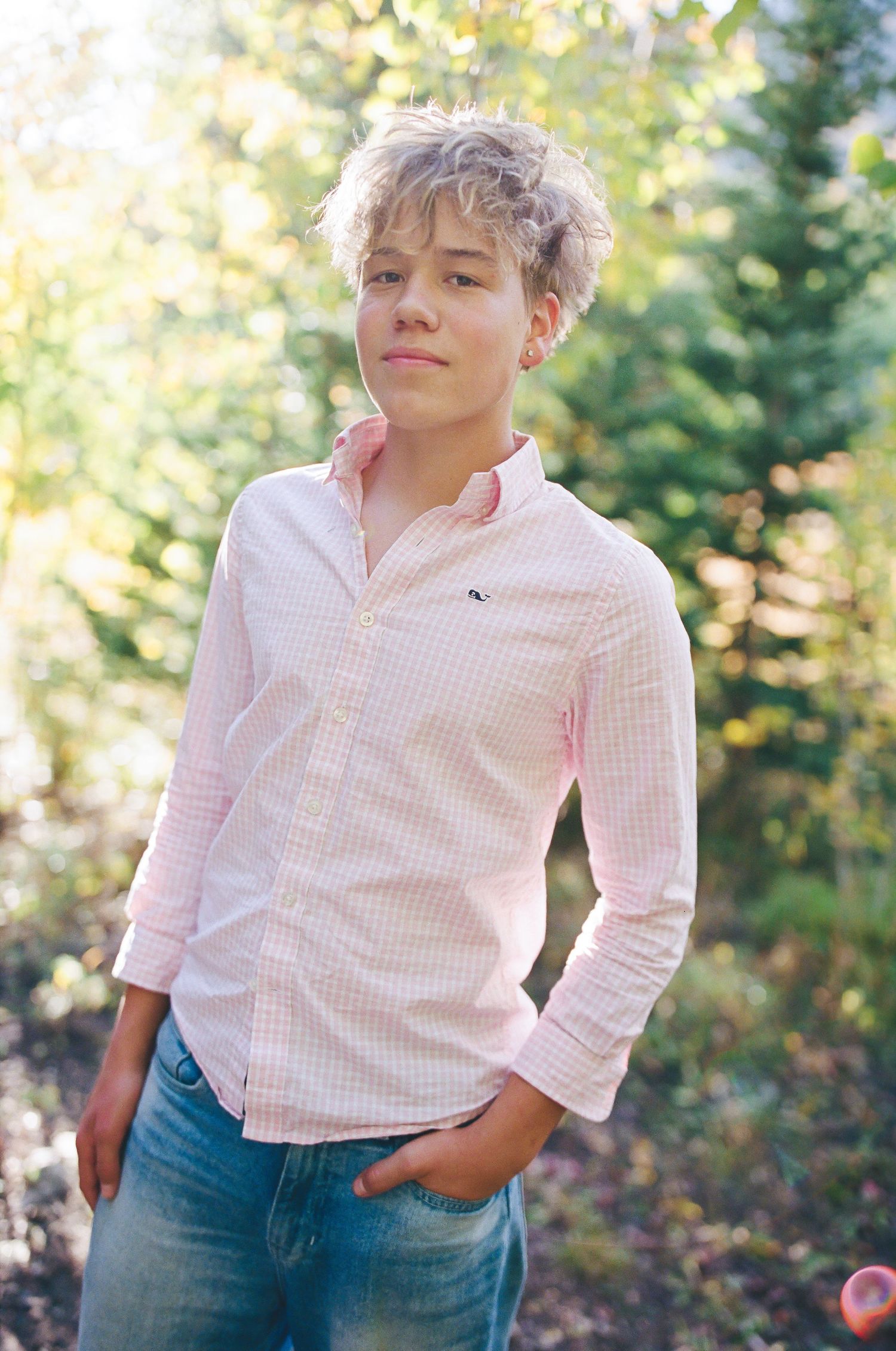 Teen boy in pink gingham shirt and jeans poses outdoors among green trees in bright sunlight.