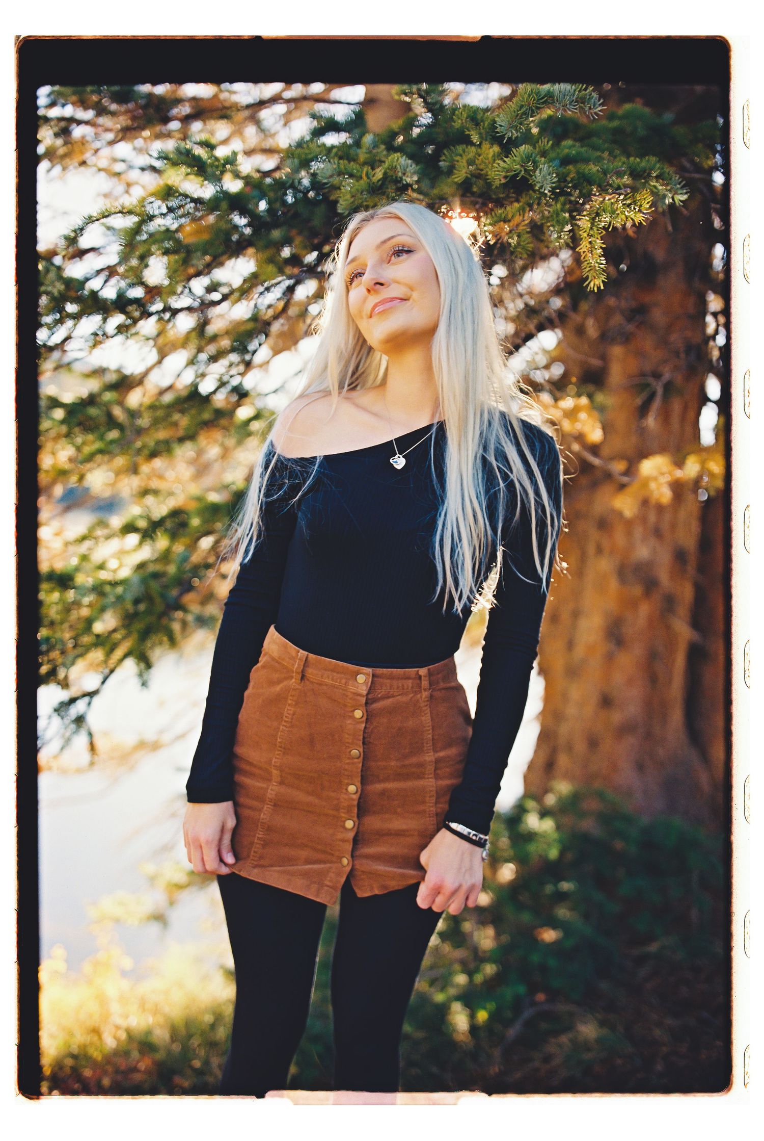 Young blonde woman in black off-shoulder top and brown corduroy skirt posing outdoors near a pine tree.