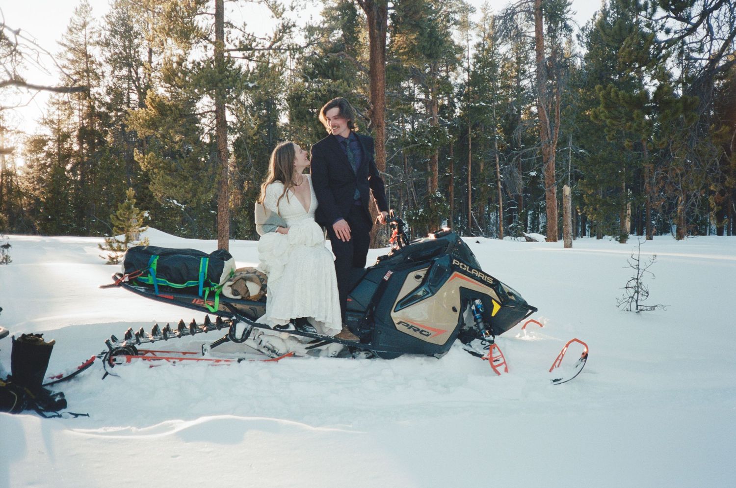 Bride and groom standing beside a snowmobile in a snowy winter forest during their outdoor wedding elopement.