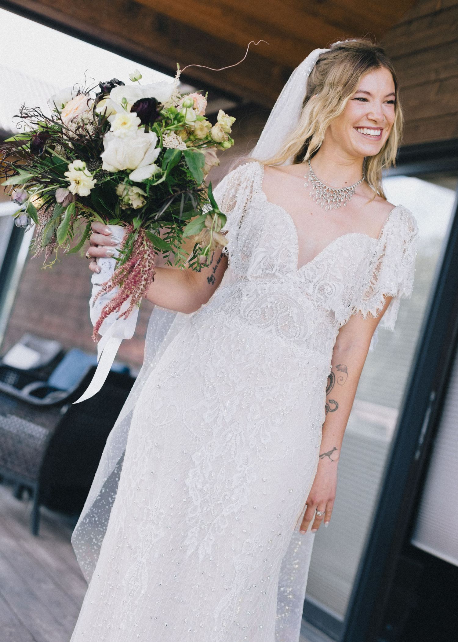Smiling bride in lace wedding dress and veil holds floral bouquet, showcasing tattoos and statement necklace.