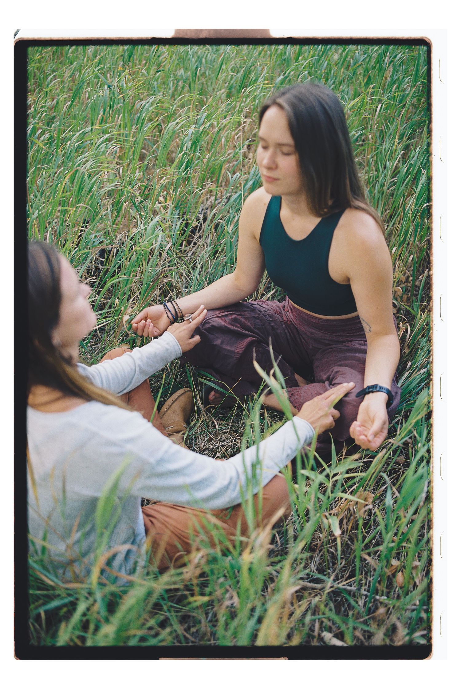 Two women sitting in tall green grass, one meditating cross-legged while the other gently holds her wrist outdoors.