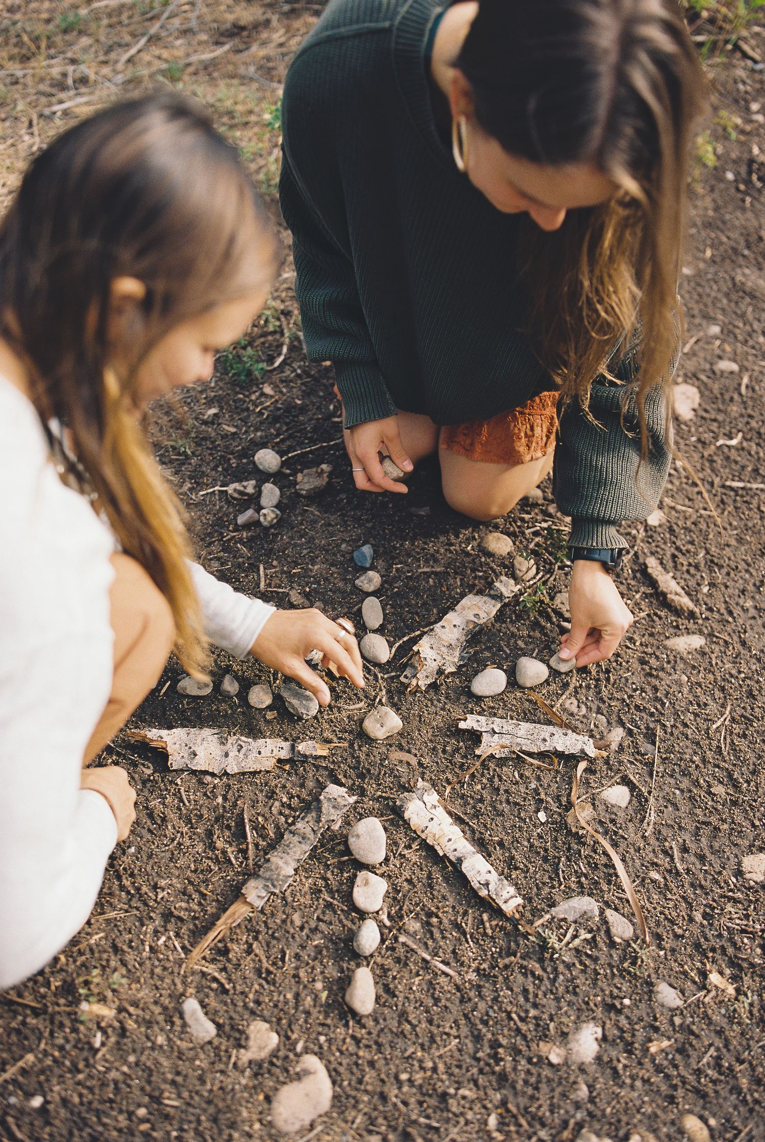 Two girls arranging rocks and sticks in a star pattern on dirt ground outdoors.