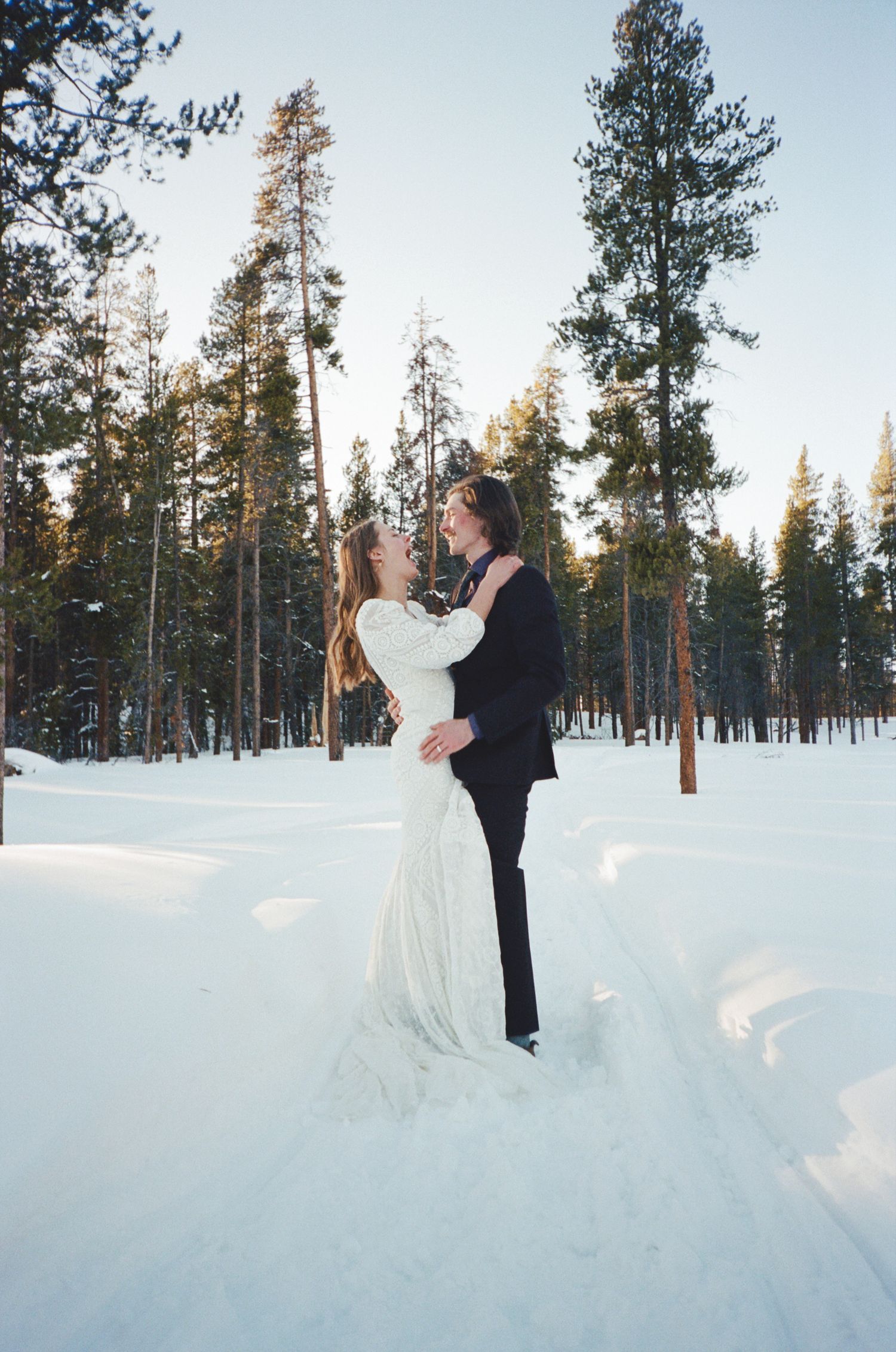 Bride and groom embrace in snowy winter forest clearing, wearing wedding attire, surrounded by tall pine trees at golden hour.