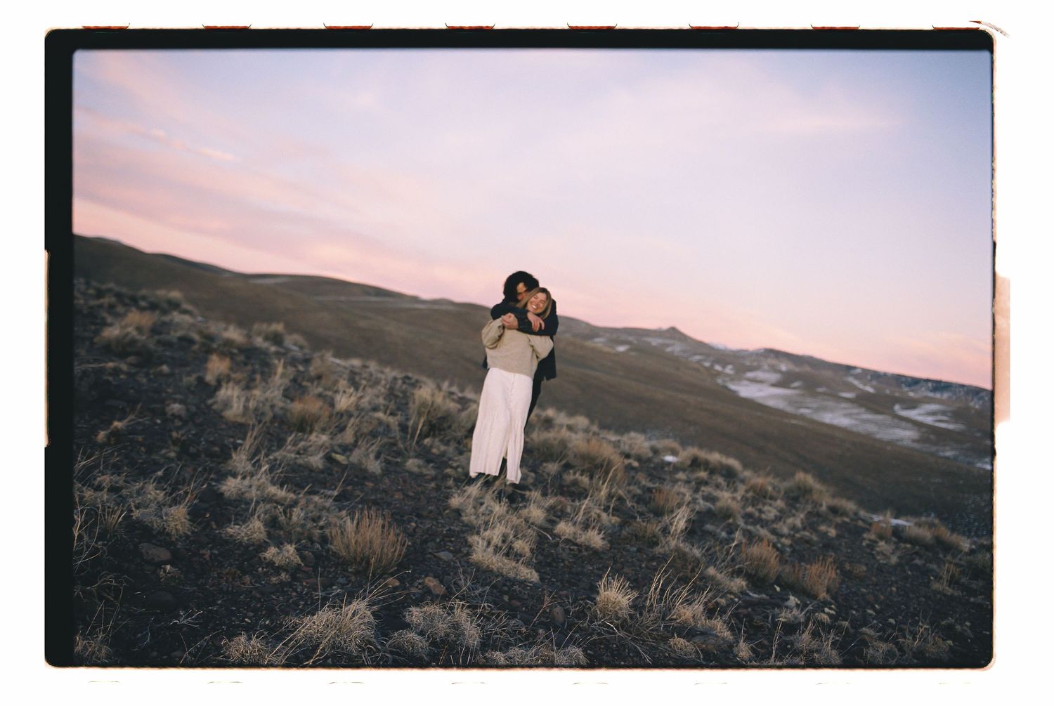 Couple embracing on a scenic hillside at sunset with a pastel sky and rolling landscape in the background.