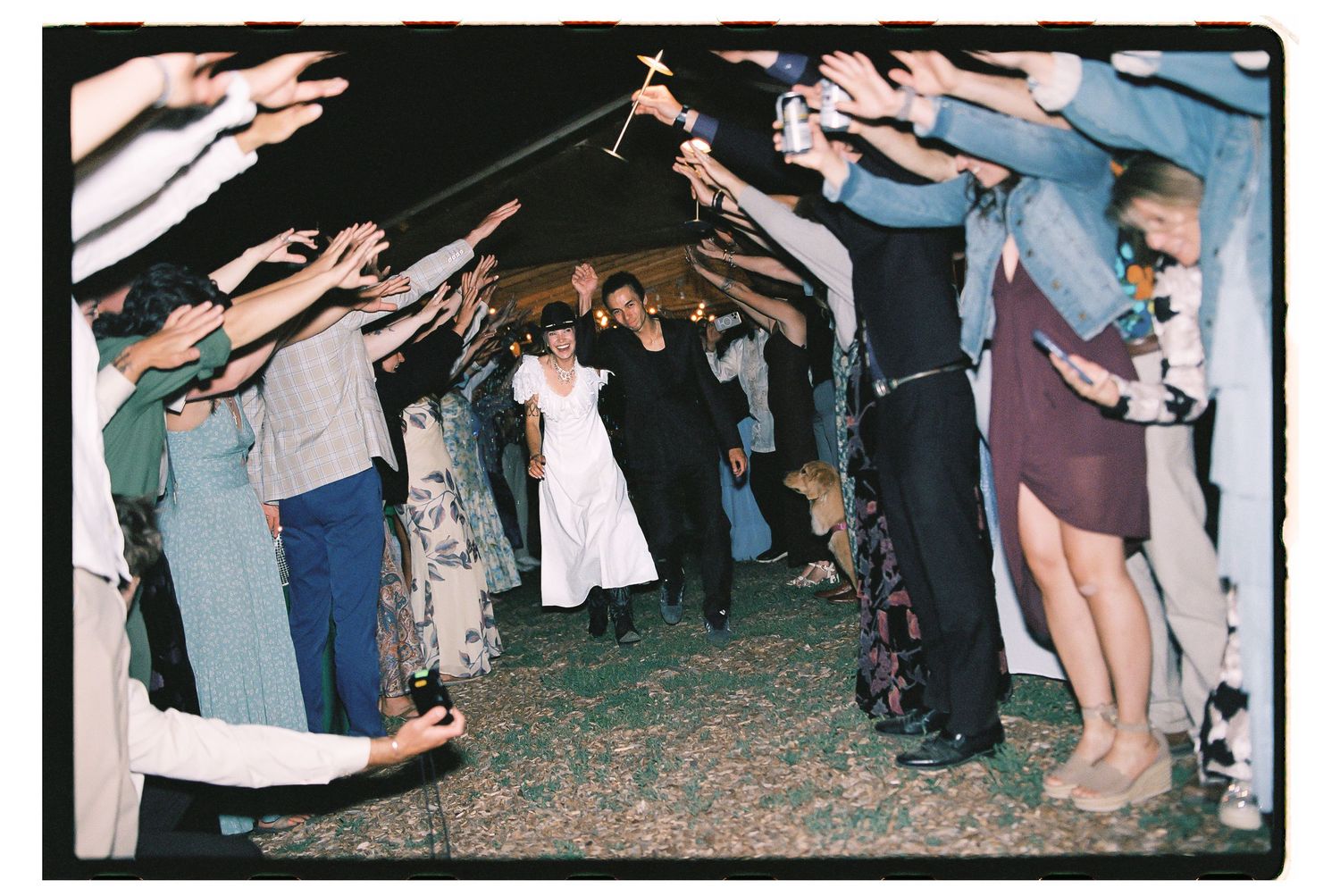 Wedding couple walks through sparkler tunnel surrounded by guests celebrating outdoors at night.