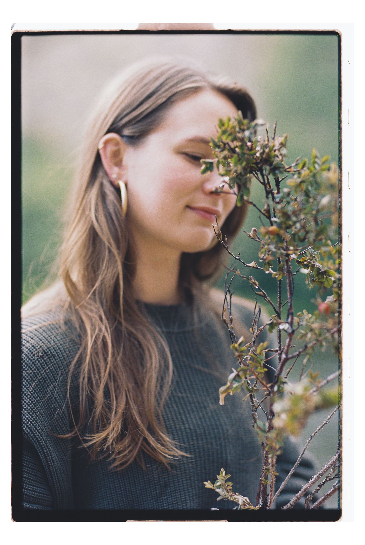 Young woman with long hair smelling a small flowering shrub outdoors, wearing a gray knit sweater and gold hoop earrings.