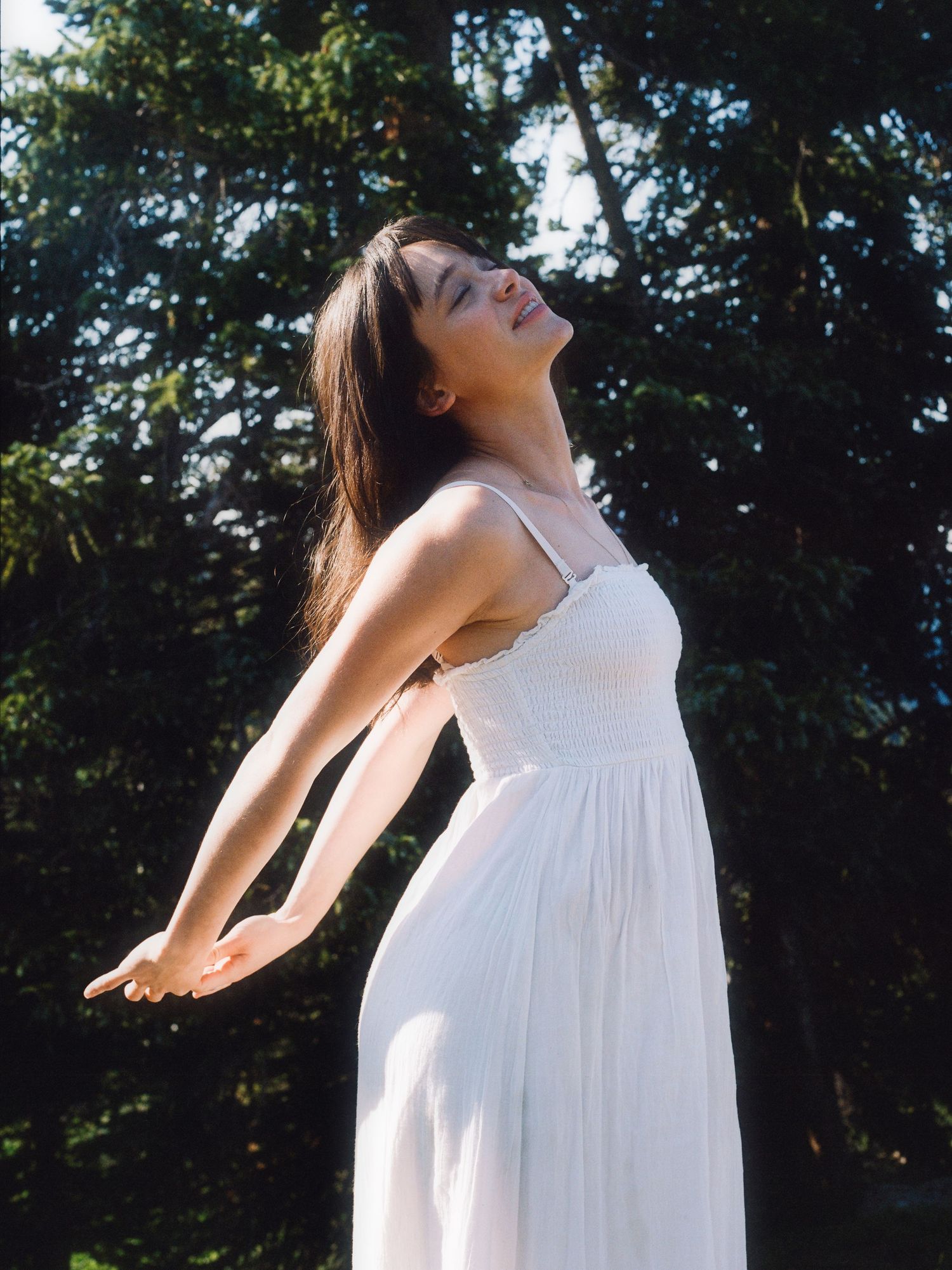 Woman in white dress with arms outstretched, eyes closed, enjoying nature among lush green trees.